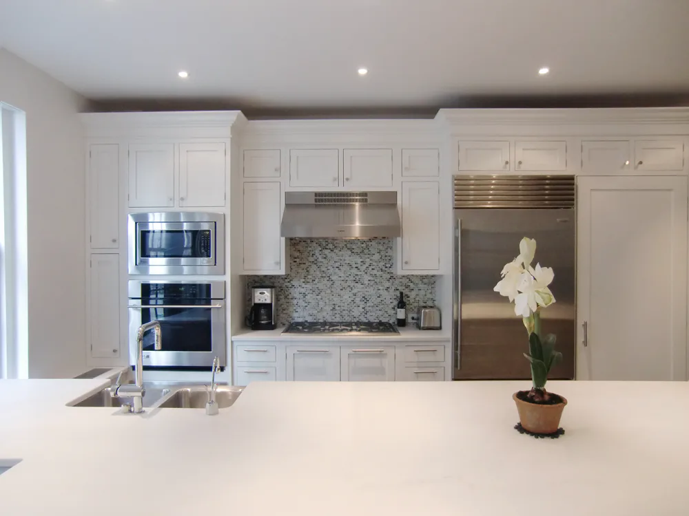 Modern white kitchen with stainless steel appliances, white cabinetry, gray mosaic backsplash, and white quartz waterfall island countertop by Arlene Avidan.