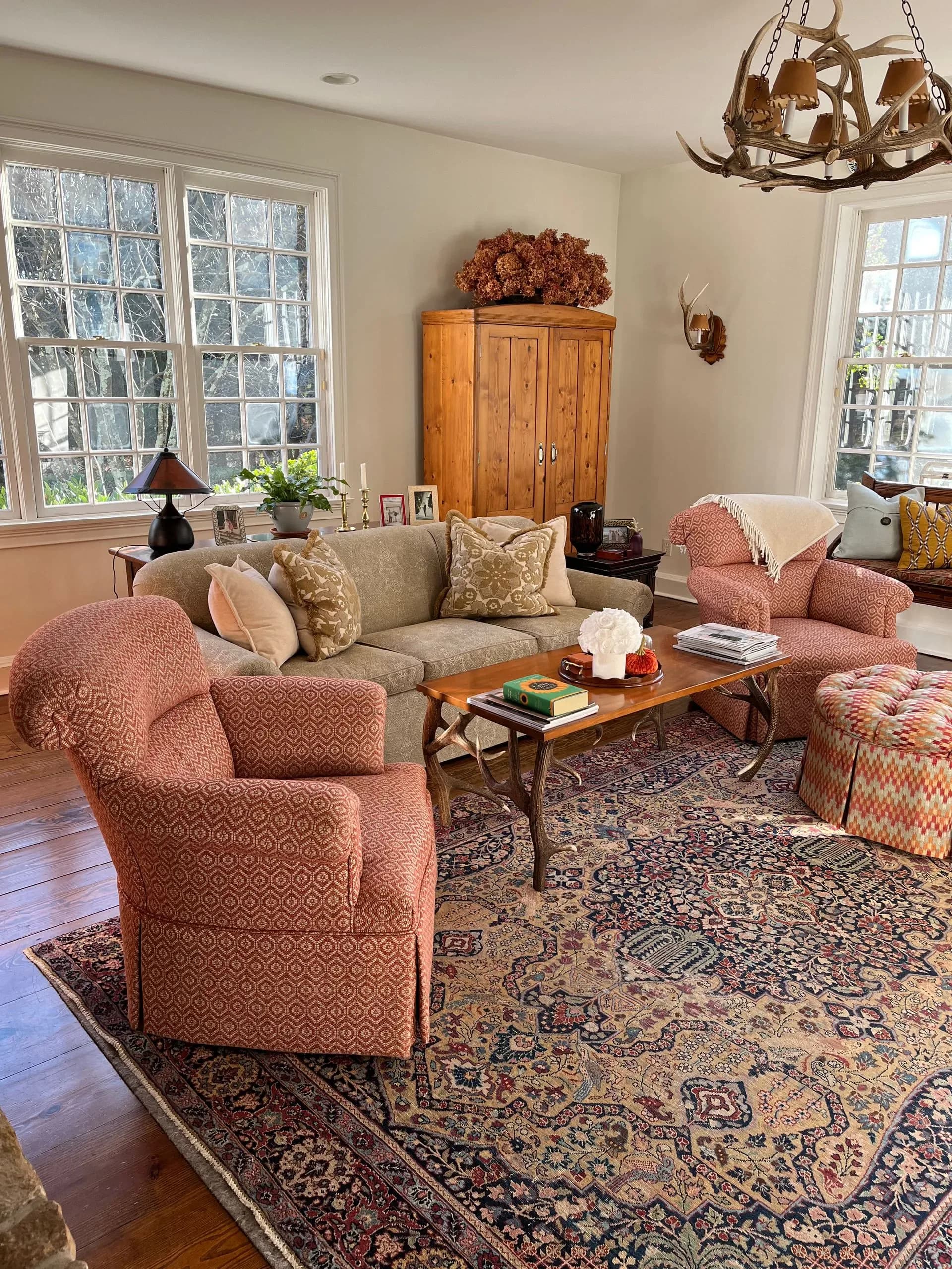 Bright living room with beige sofa, coral accent chairs, patterned area rug, wooden coffee table, and antler chandelier by Arlene Reilly.