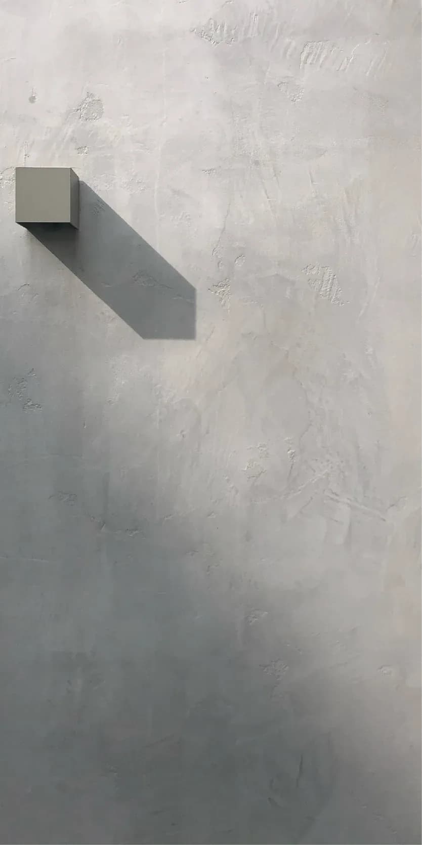 Minimalist interior detail showing a geometric gray cube-shaped wall element casting a sharp shadow on a textured white plaster wall by Arman Safavi.