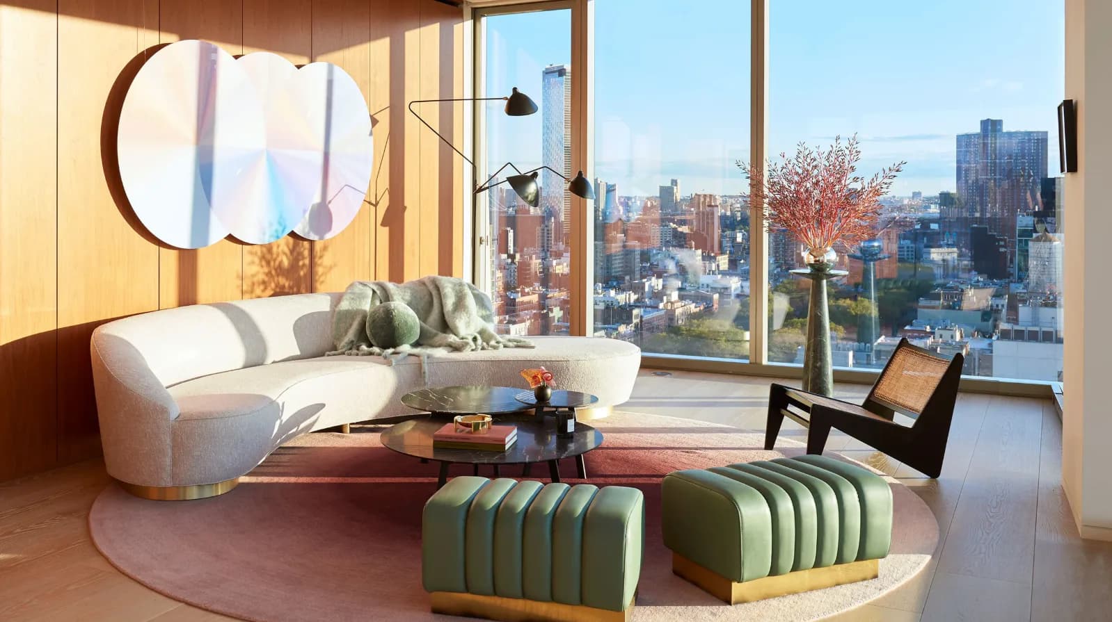 Modern high-rise apartment with curved cream sofa, green striped ottomans, circular mirror, and expansive city skyline views by Armann Ortega.
