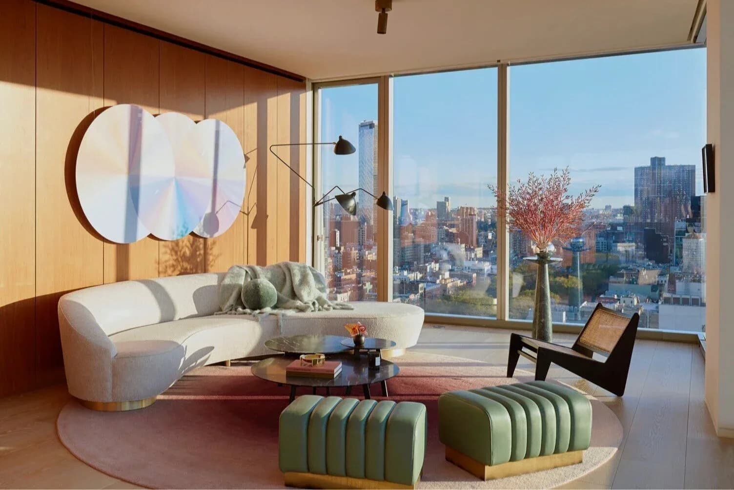 Modern high-rise apartment with curved cream sofa, green ribbed ottomans, round mirror, and expansive city skyline views through floor-to-ceiling windows by Armann Ortega.