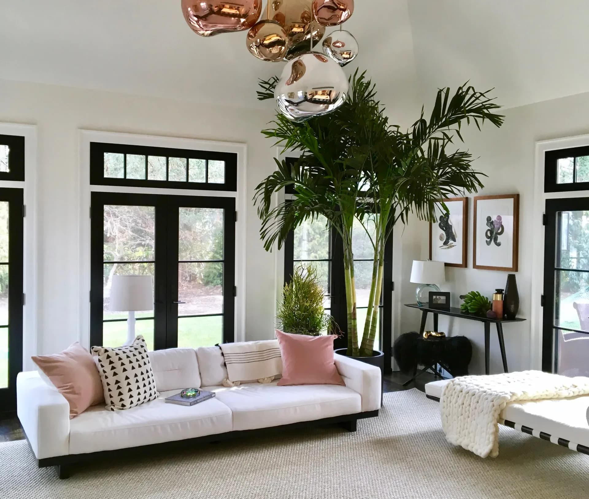 Bright modern living room with cream sofa, tall potted plants, copper pendant lights, black-framed windows, and neutral palette by Armann Ortega.