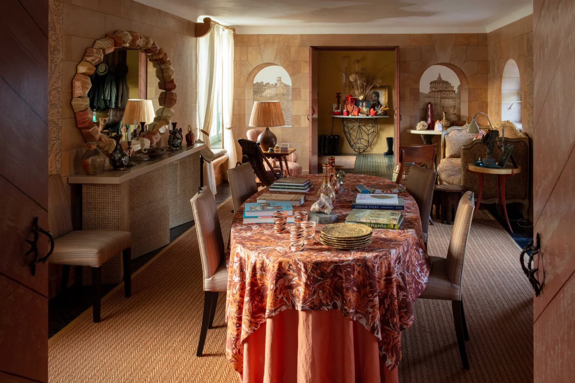 Warm terracotta dining room with patterned red tablecloth, arched doorways, period furnishings, and ornamental mirrors, designed by Ashley Hicks.