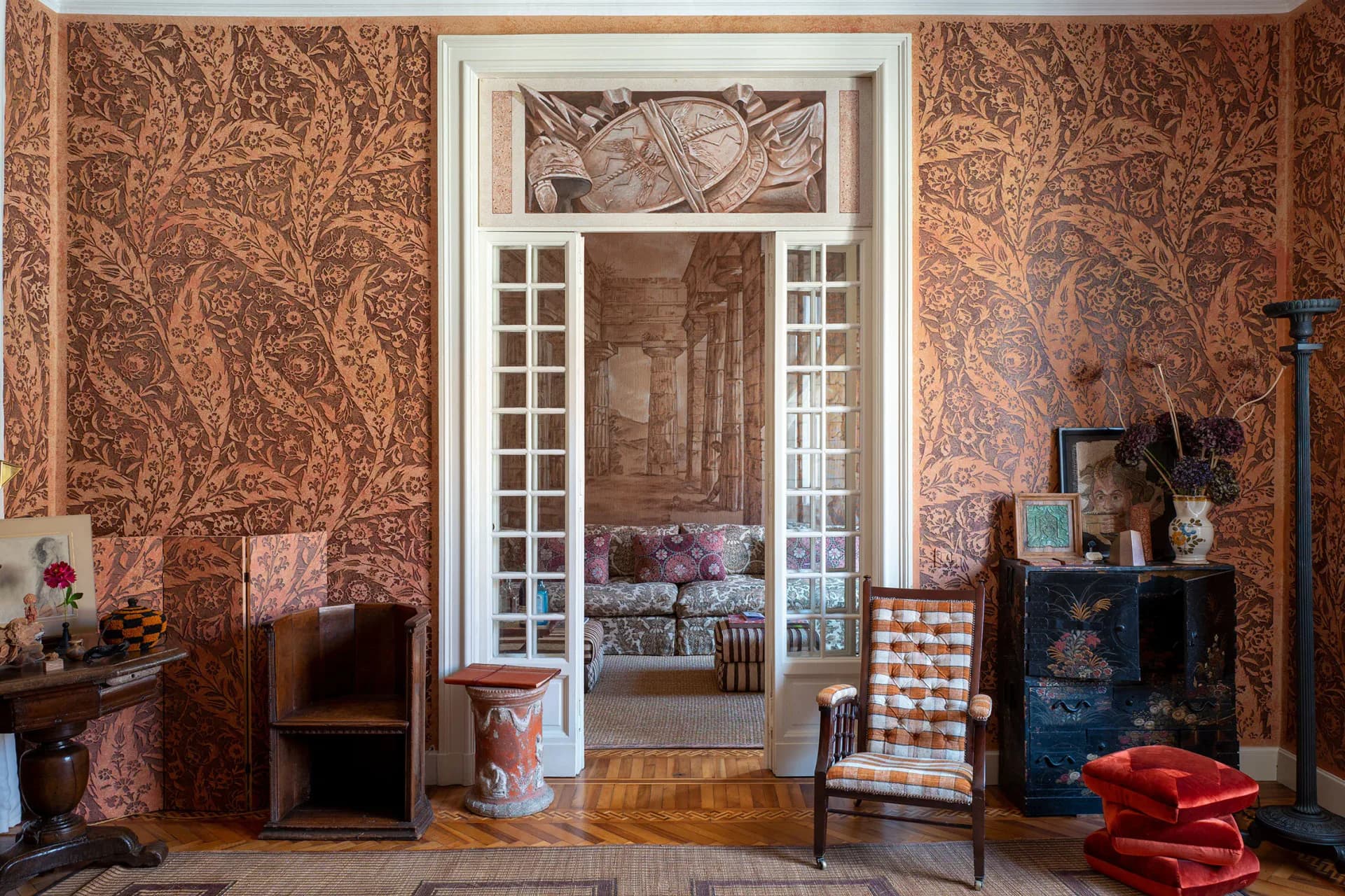 Ornate living room with rust-colored damask wallpaper, white French doors, wooden parquet flooring, and period furnishings by Ashley Hicks.