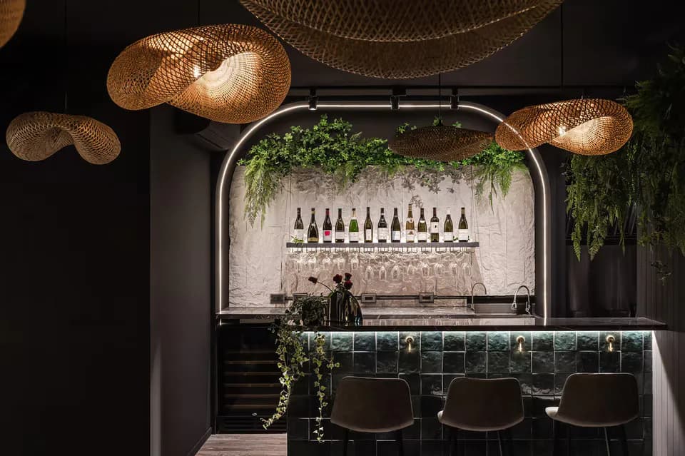 Modern bar interior with dark teal tile backdrop, suspended wine bottles, woven pendant lights, living green wall, and black bar seating by Ashley Liu.