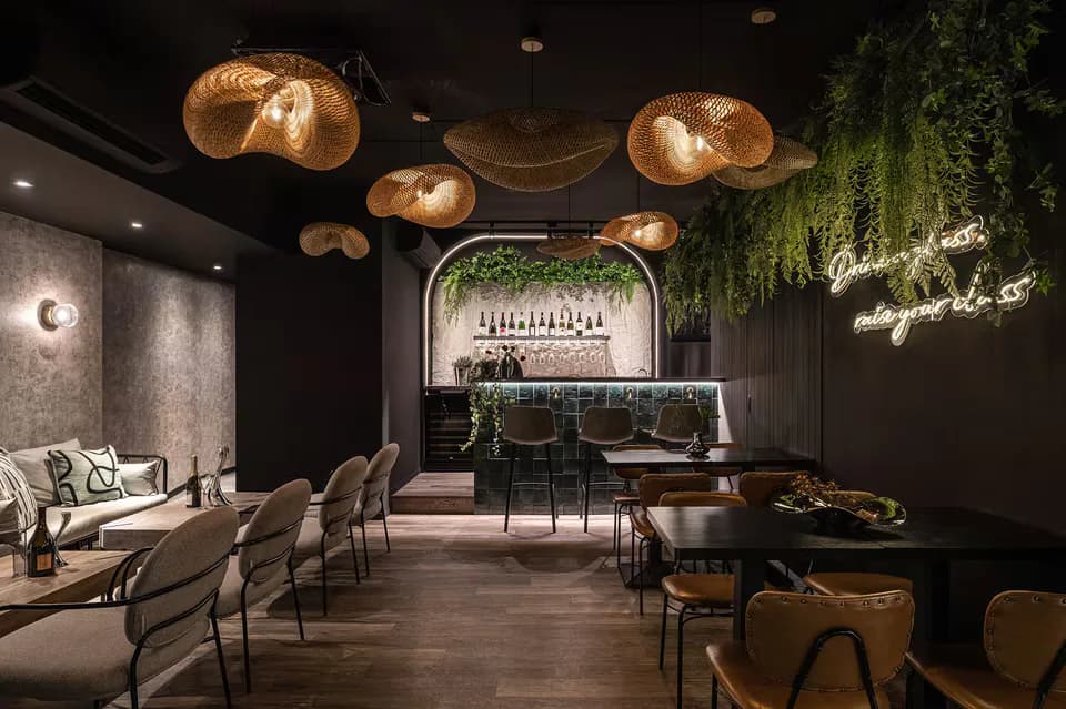 Dark luxurious lounge interior with copper pendant lighting, green plant installations, a bar counter, seating areas, and wooden flooring, designed by Ashley Liu.