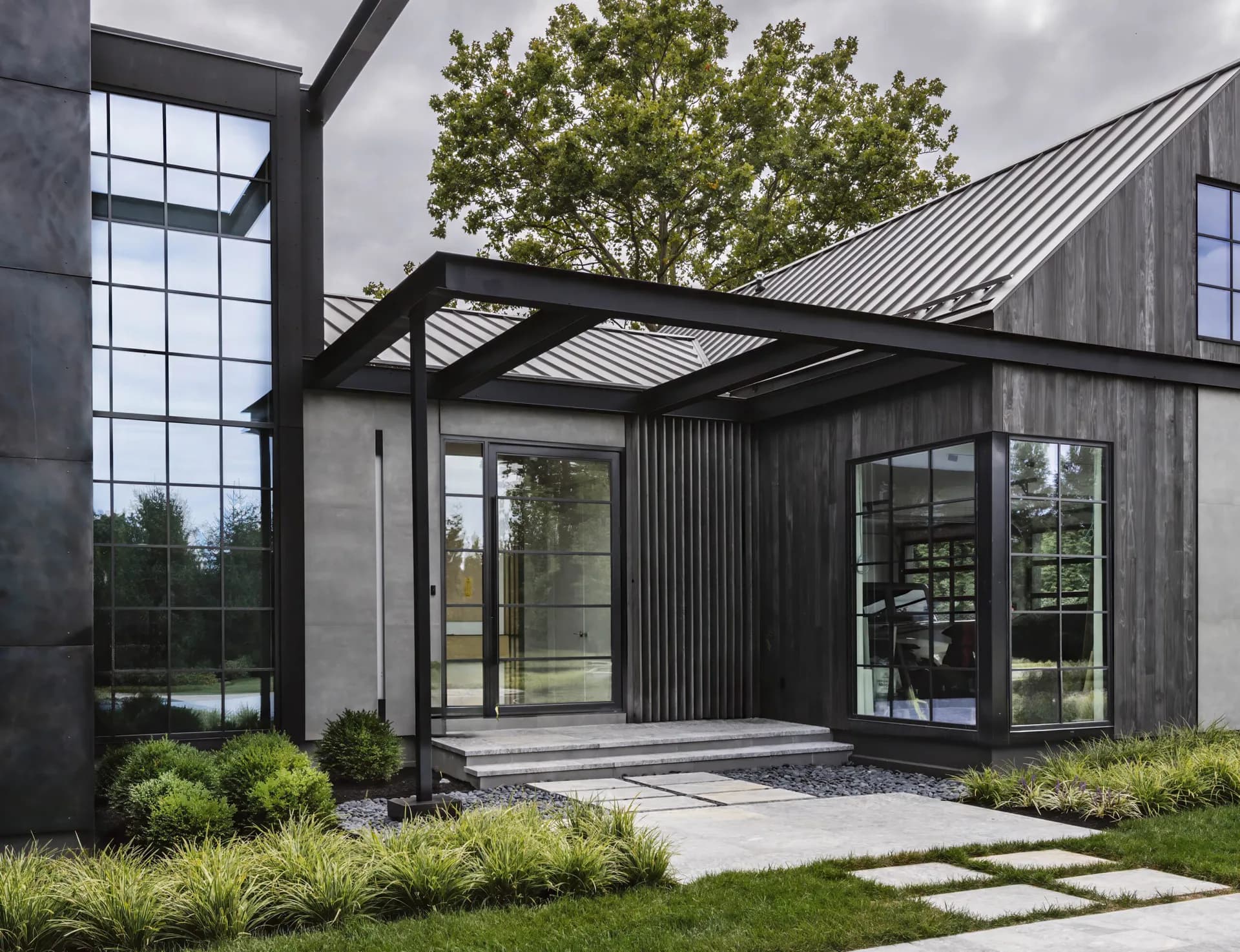 Modern black-metal pergola structure with glass doors and windows overlooking landscaped grounds with ornamental grasses by Ashley Rose Walsh.