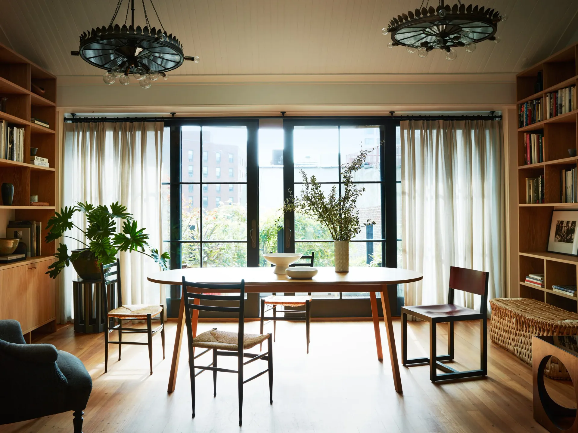 Bright study workspace with wood desk, mid-century chairs, floor-to-ceiling windows, floating shelving, and pendant lighting by Ashok Childs.