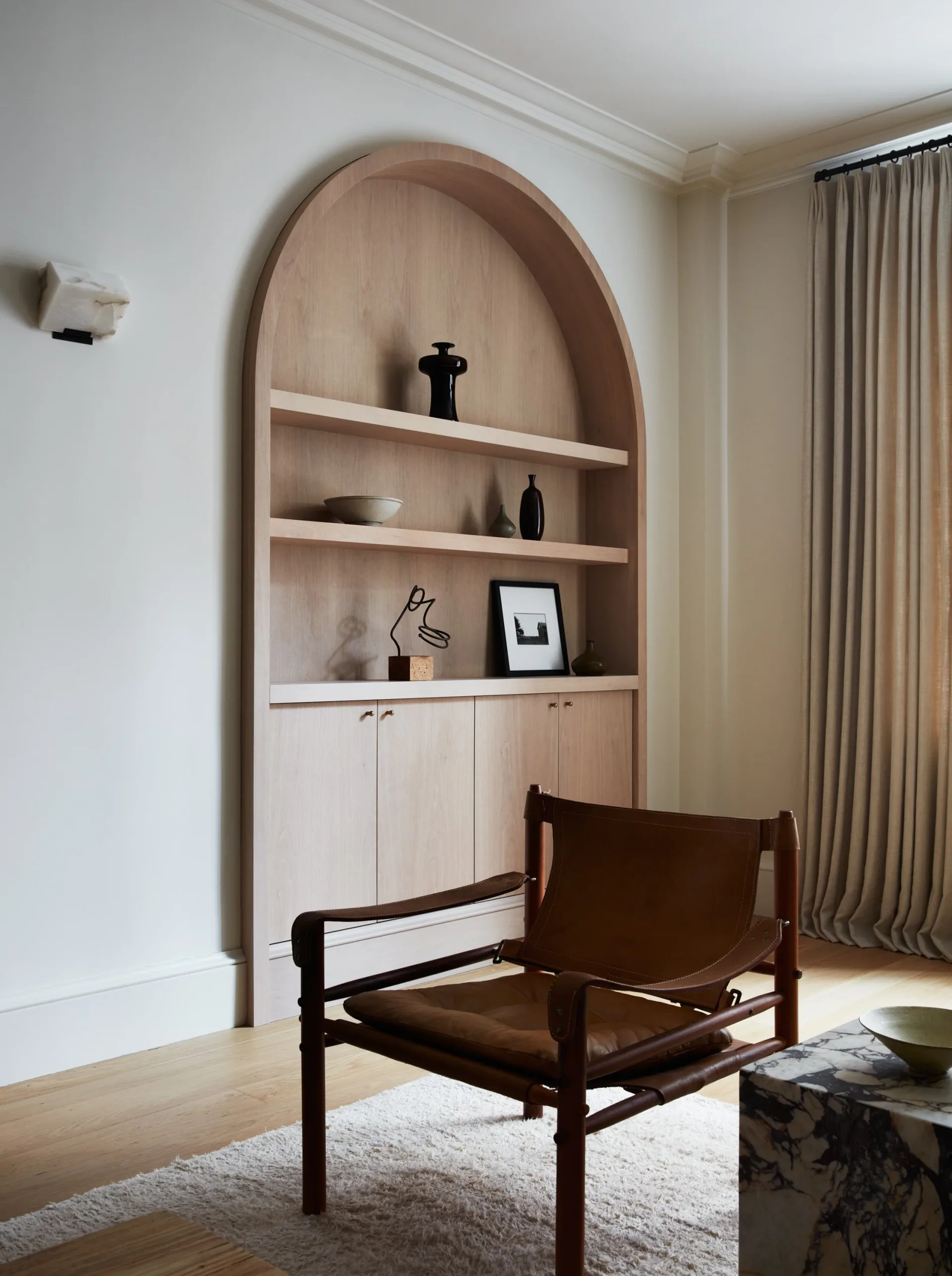 Minimalist interior featuring a curved arched bookcase in light wood with open shelving and lower cabinetry, paired with a dark wooden mid-century chair, by Ashok Childs.
