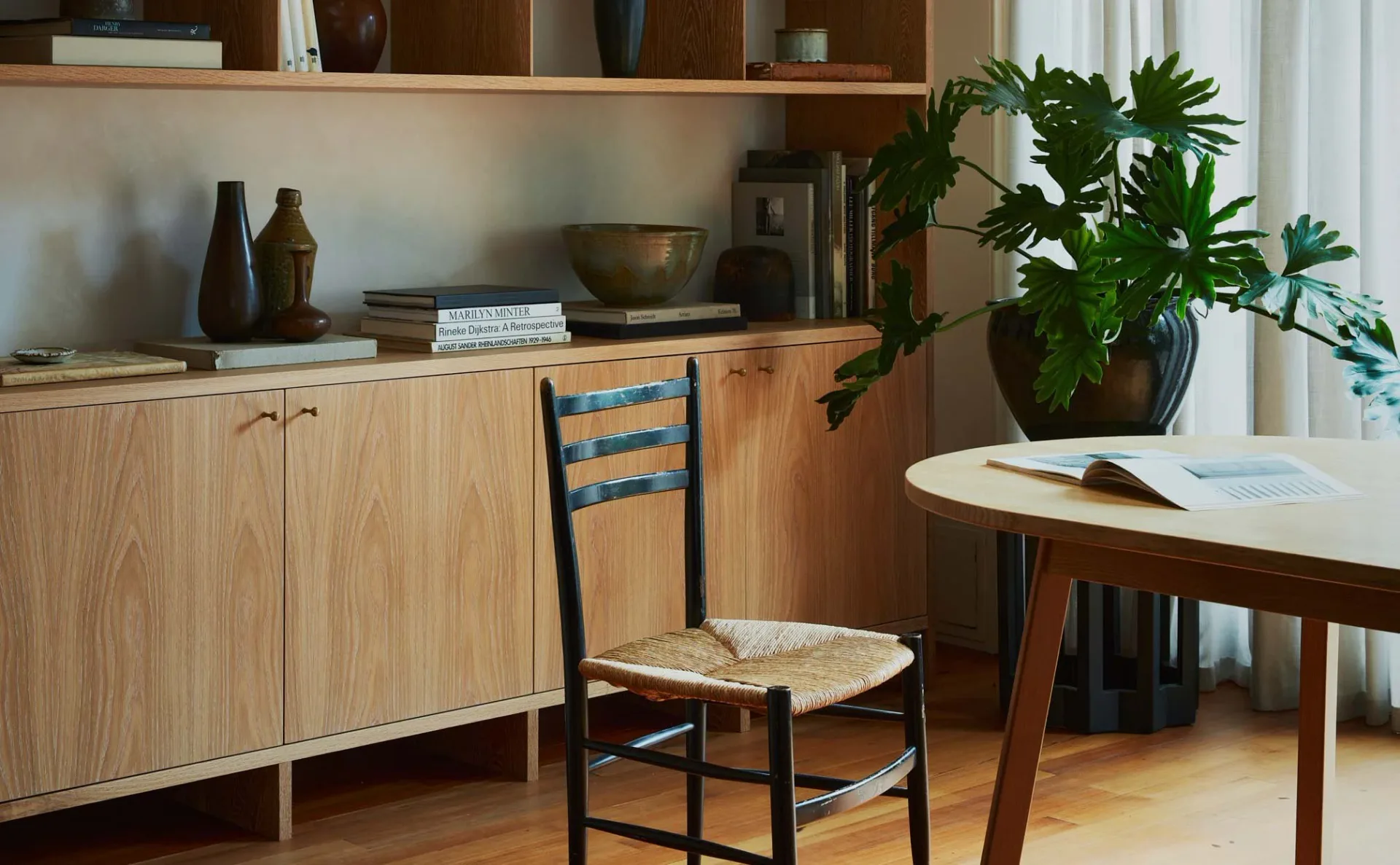 Warm wood cabinetry and shelving with natural materials, black ladder-back chair, potted plant, and round dining table in a minimalist interior by Ashok Childs.