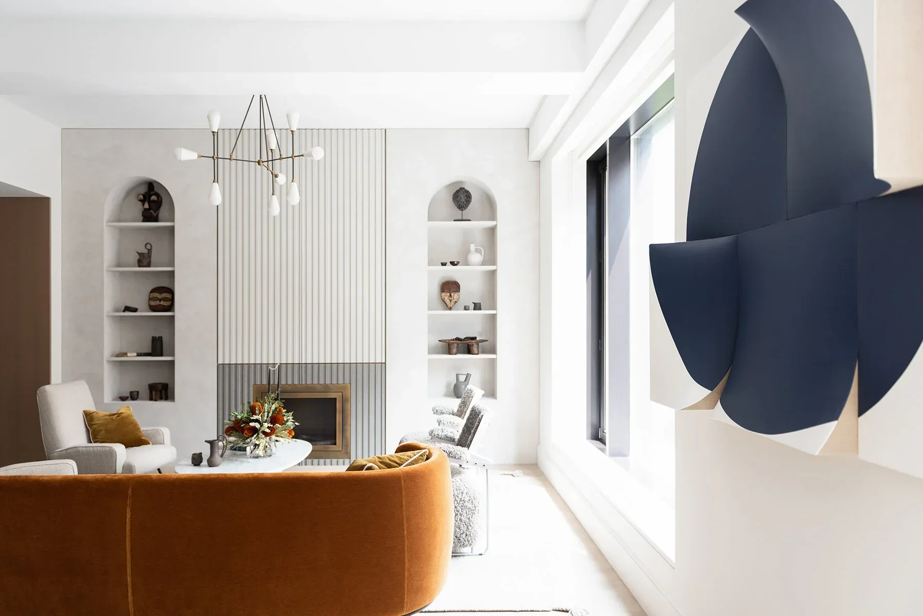 Bright modern living room with caramel leather sofa, white built-in shelving, fireplace, geometric wall art, and large windows by Ashok Childs.