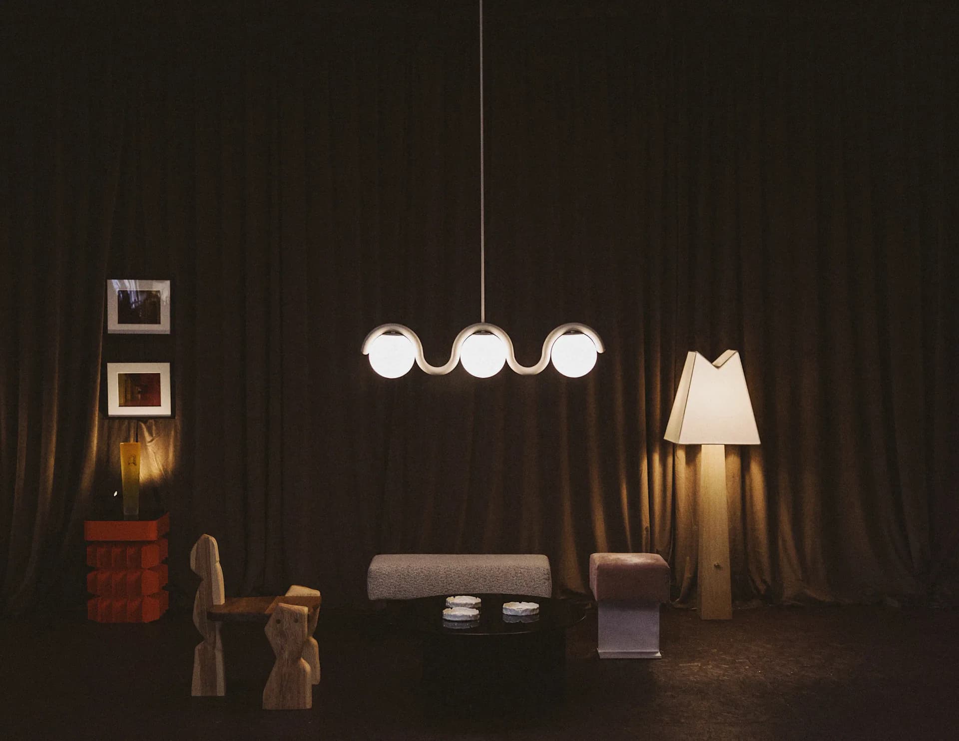 Dark interior space with draped curtains, featuring a sculptural pendant light with three connected spheres, upholstered seating, wooden furniture pieces, and warm accent lighting by Astraeus Clarke.