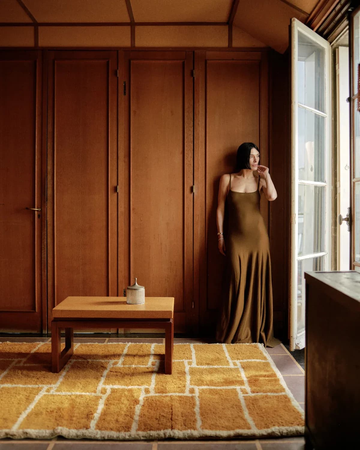 Warm terracotta interior with floor-to-ceiling wooden cabinetry, geometric patterned rug, and woman in olive dress by Athena Calderone.