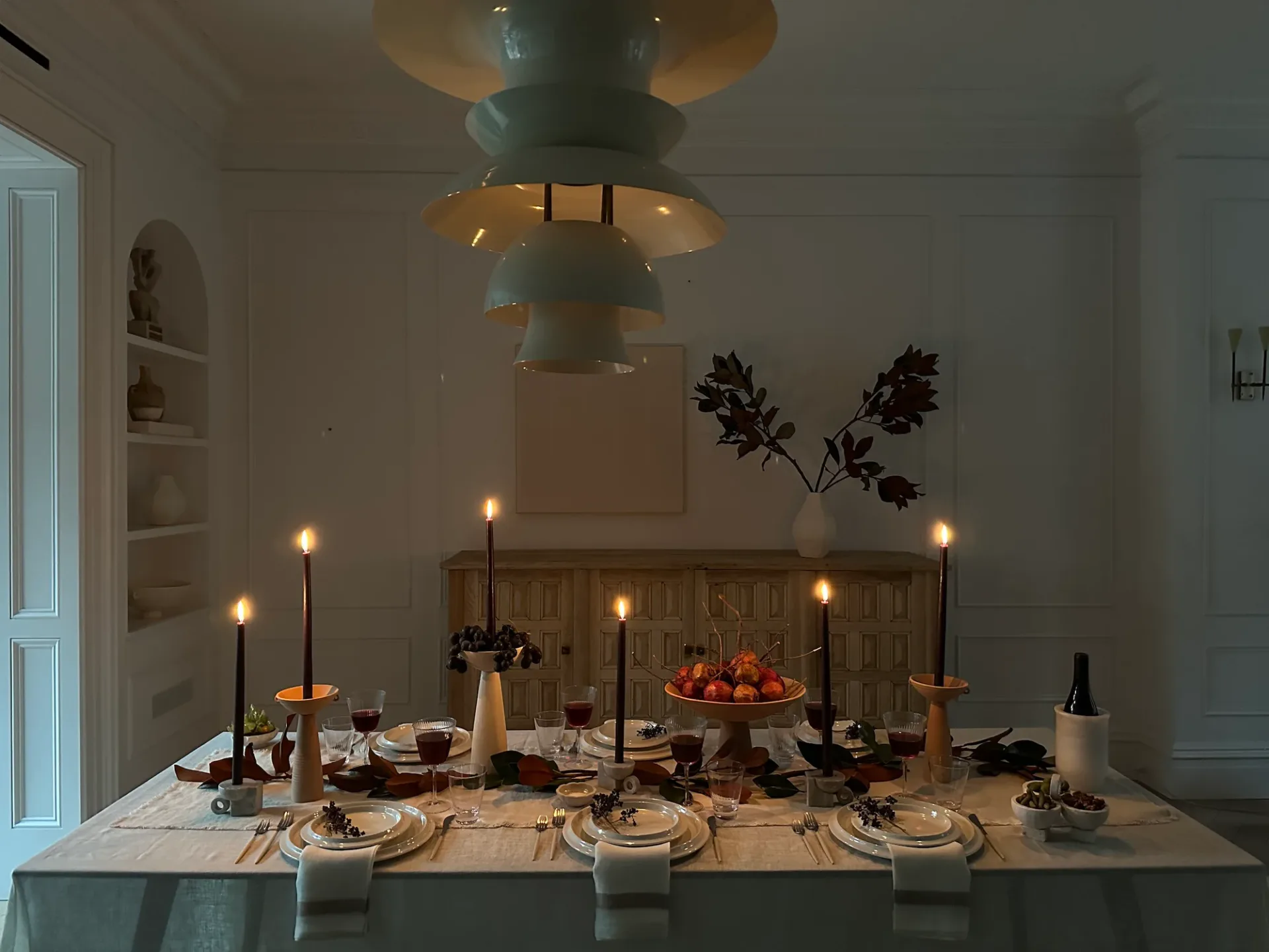 Formal dining table setting with tiered brass pendant lighting, candlelit place settings, dried florals, and autumn fruit centerpiece by Athena Calderone.