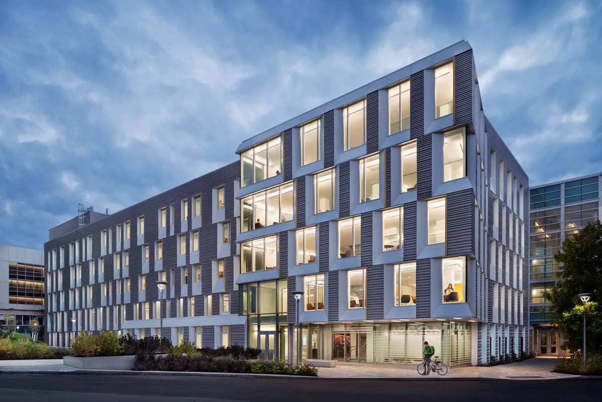 Modern four-story residential building with dark facade, illuminated rectangular window grid, ground-level glass storefront, and evening sky backdrop by Attila Uysal.