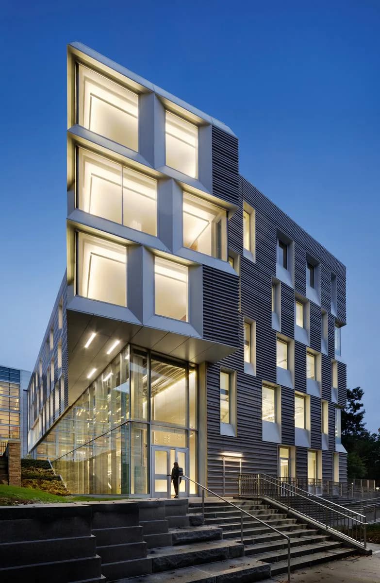 Modern residential building with illuminated facade featuring golden-framed windows, dark horizontal louvers, and a ground-level glass entrance with stairs, photographed at dusk by Attila Uysal.