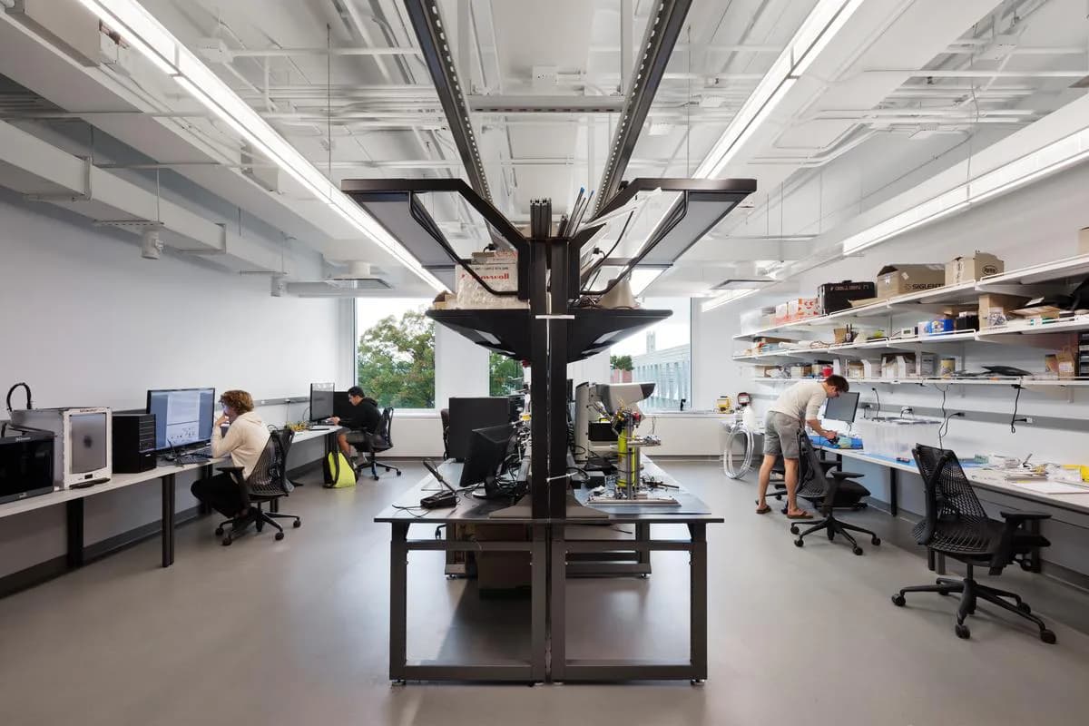 Modern laboratory or research workspace with central black structural column, workstations with computers and equipment, bright fluorescent ceiling lights, and open floor plan with shelving, designed by Attila Uysal.