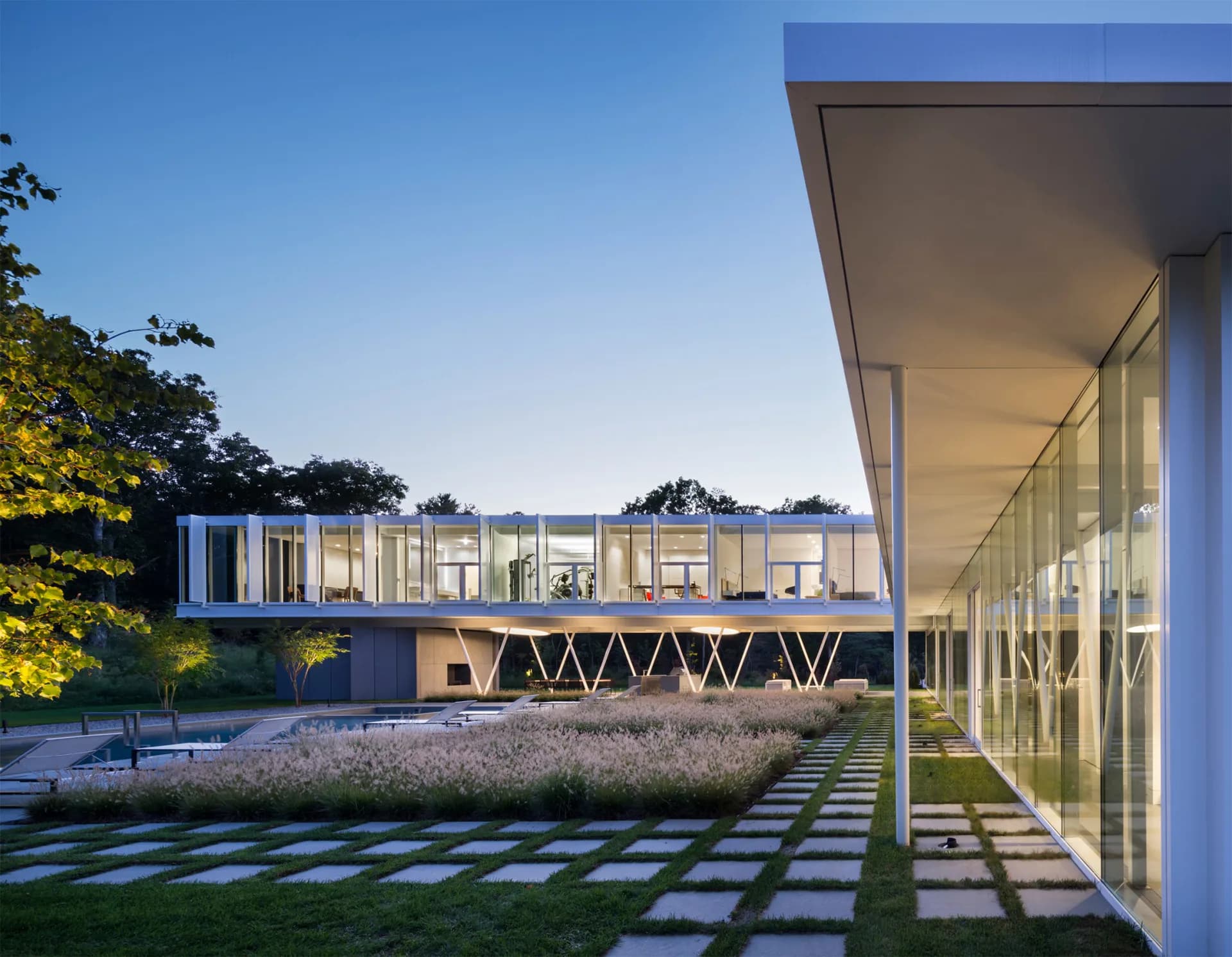 Modern residential architecture with cantilevered deck, extensive glazing, and geometric paving patterns, illuminated at dusk by Audrey Matlock.
