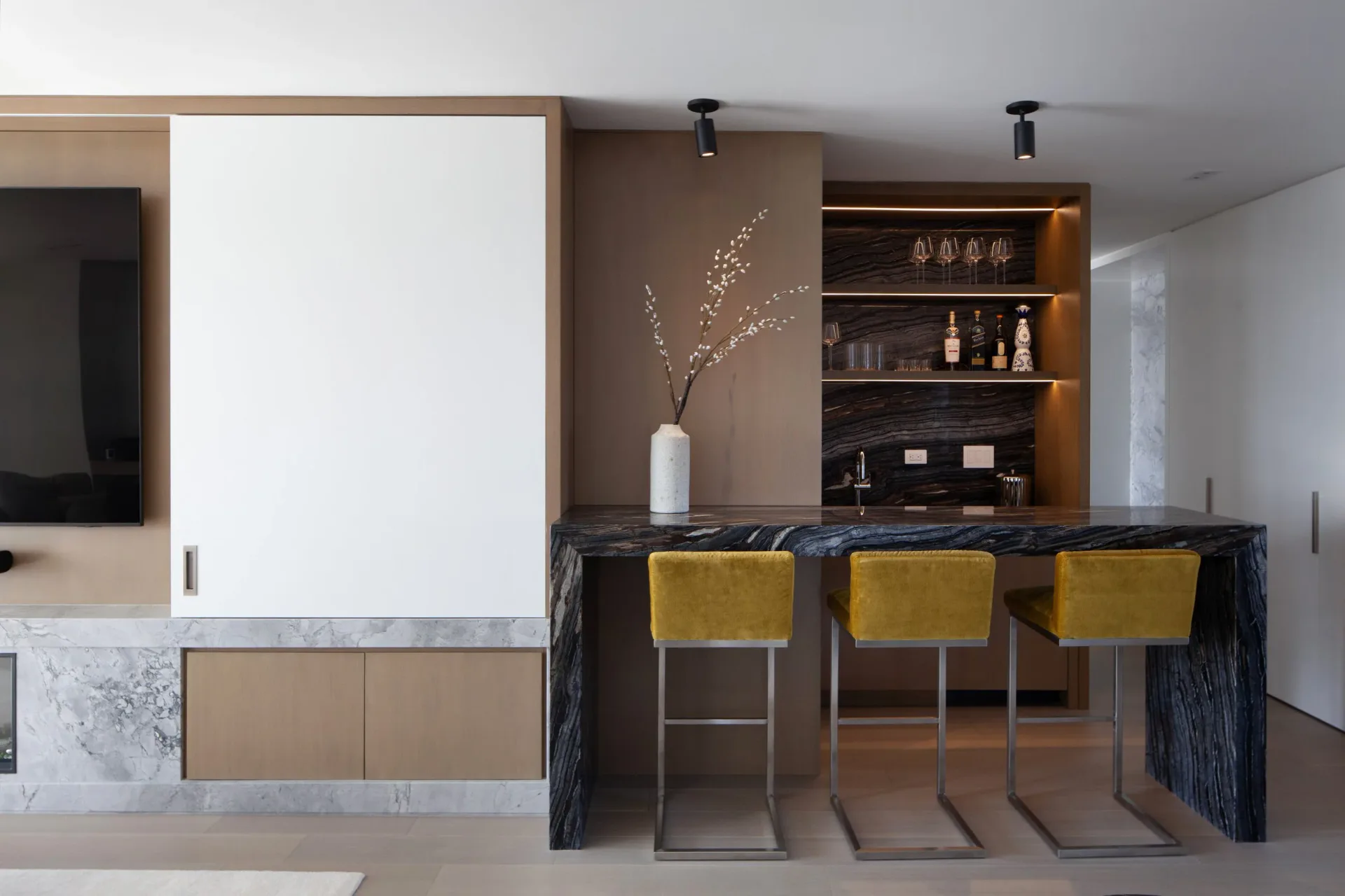 Modern kitchen and bar interior with dark marble island, mustard velvet stools, built-in shelving, and minimalist finishes by August Liau.