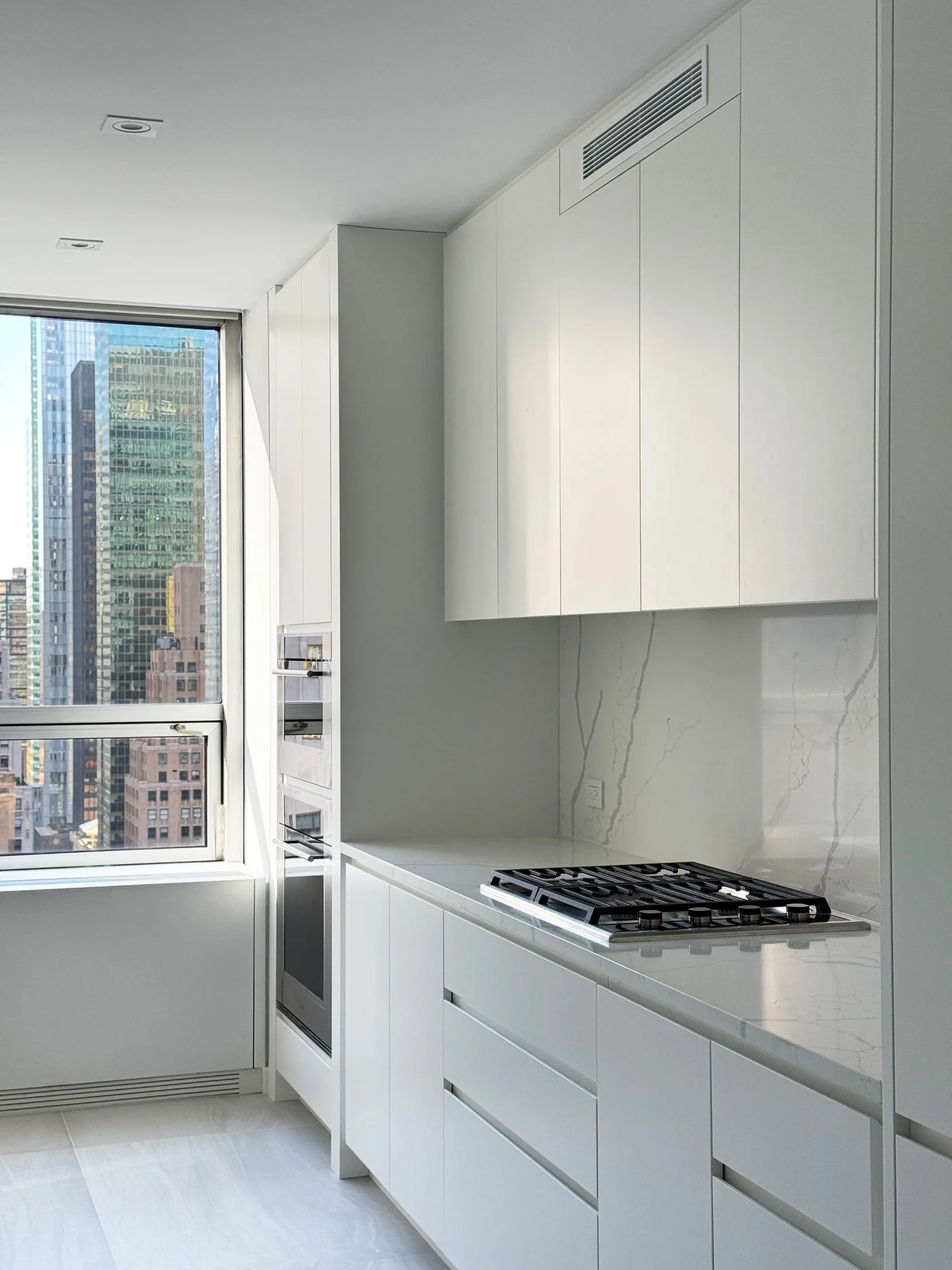 Minimalist white kitchen with marble backsplash, gas cooktop, and floor-to-ceiling cabinetry overlooking a New York City skyline by August Liau.