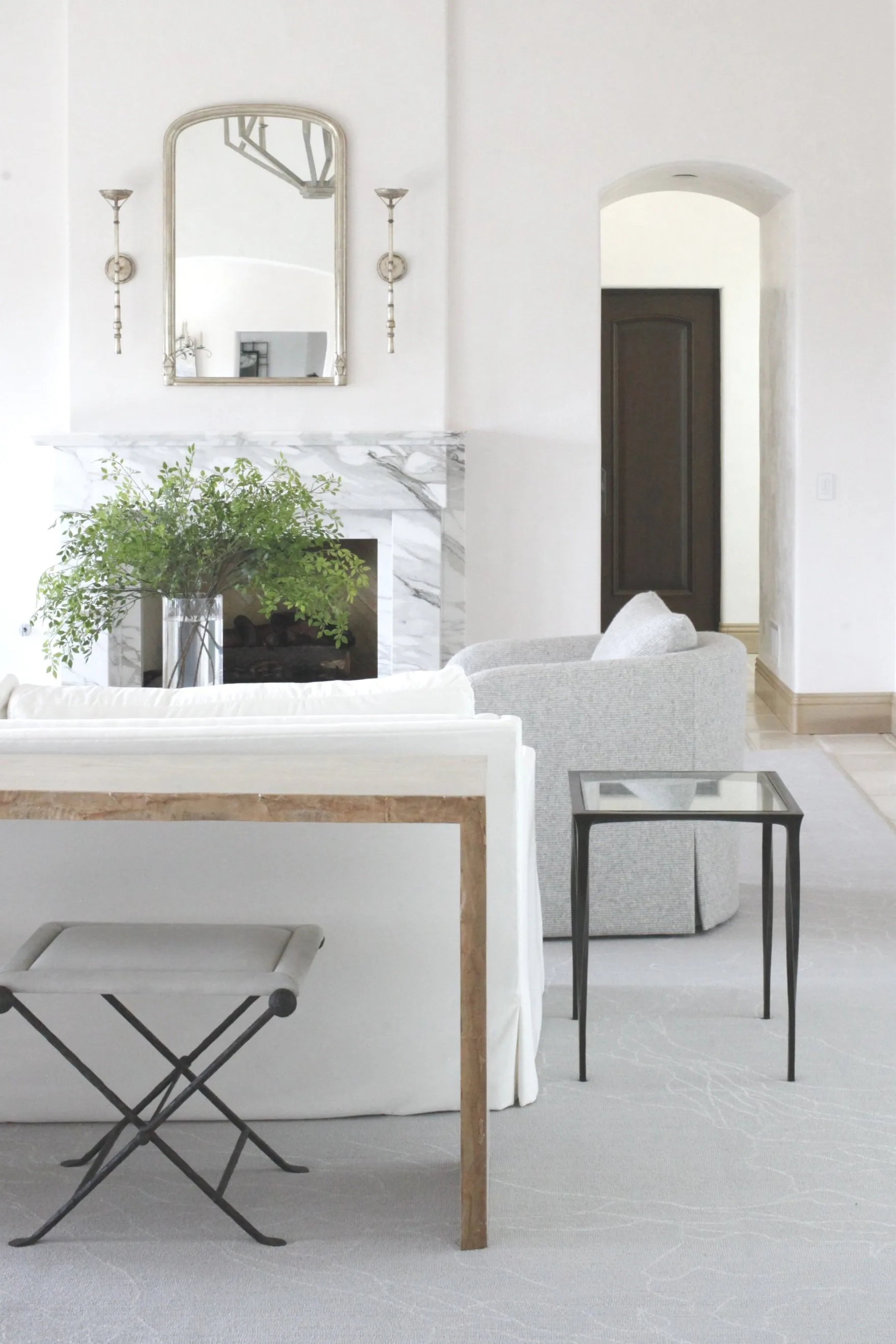 Minimalist bathroom with marble countertop, arched mirror with sconces, white cabinetry, potted greenery, and metal stools by Ava Lunder.