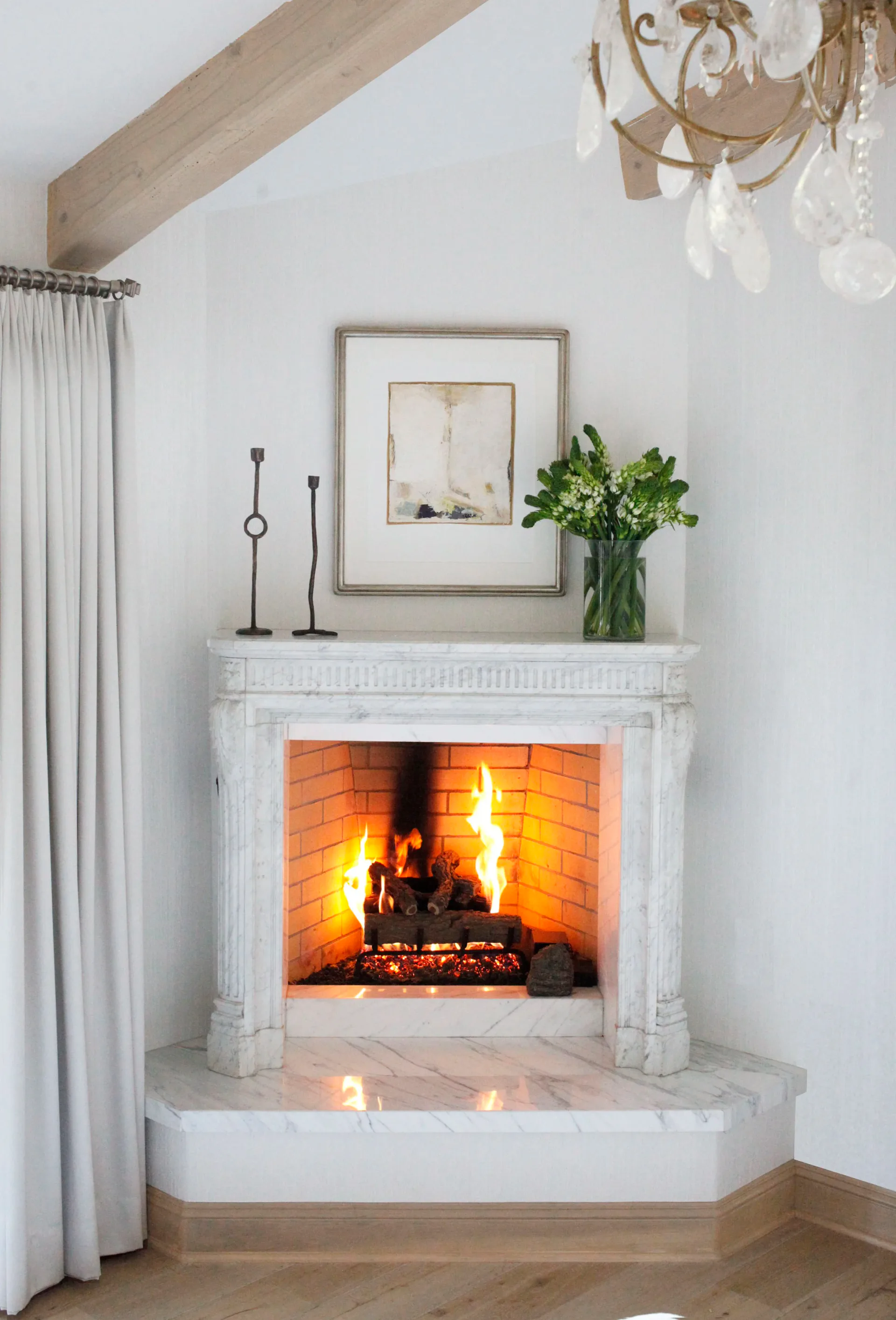 A white fireplace with marble hearth and ornate mantelpiece features a roaring fire, styled with framed artwork, candles, and fresh greenery by Ava Lunder.