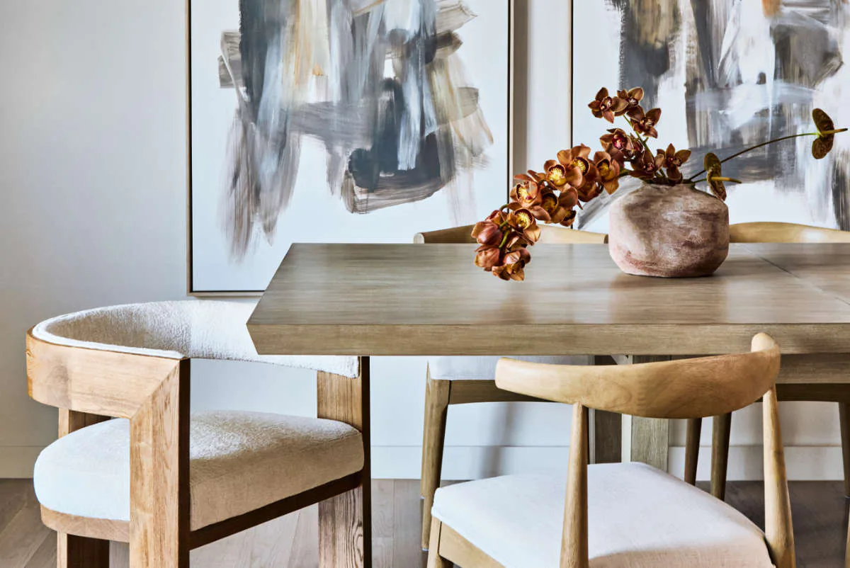 Modern dining room with natural wood table, cream upholstered chairs, dried botanicals in ceramic vessel, and abstract artwork by Avery Johnson.