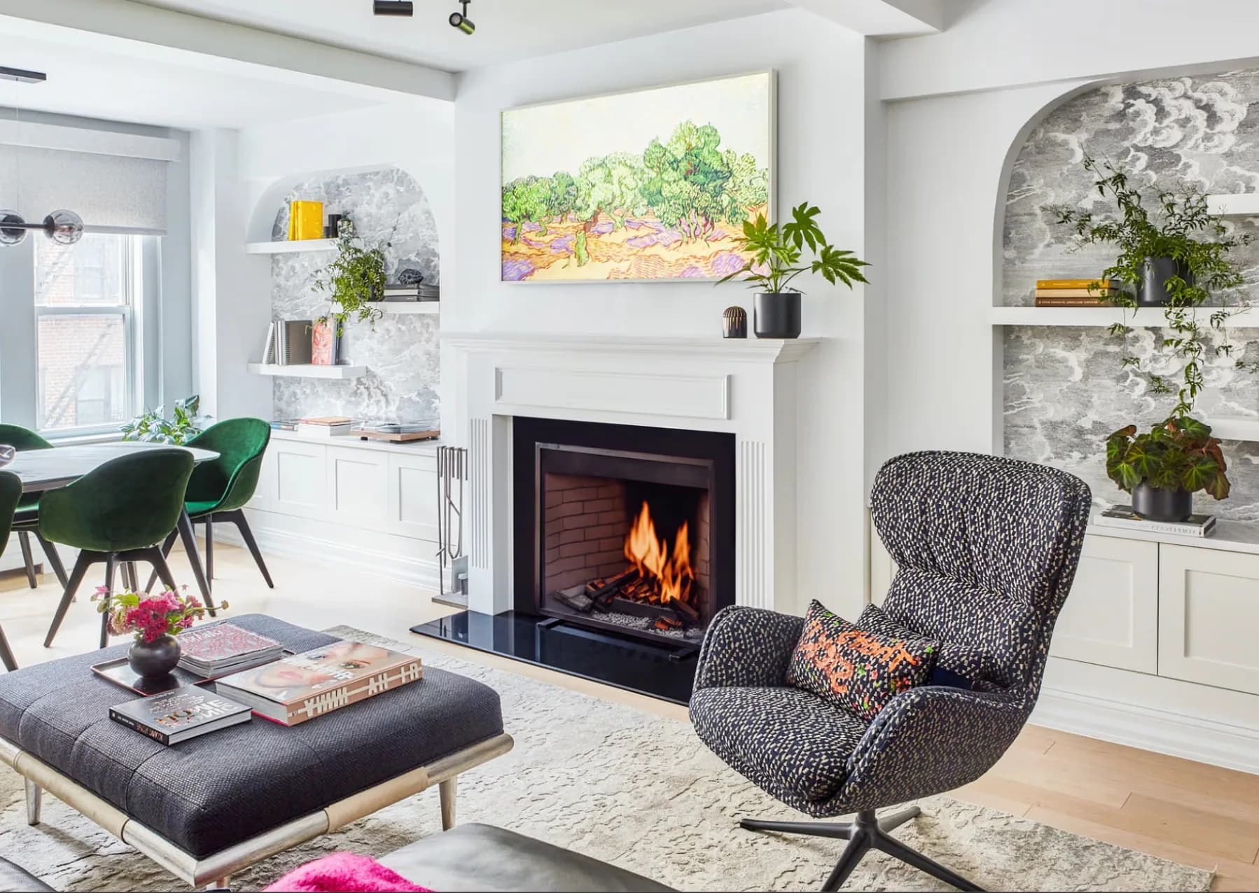Modern living room with fireplace, gray armchair, black ottoman, white built-in shelving, green accent chairs, and warm wood flooring by Avery Johnson.