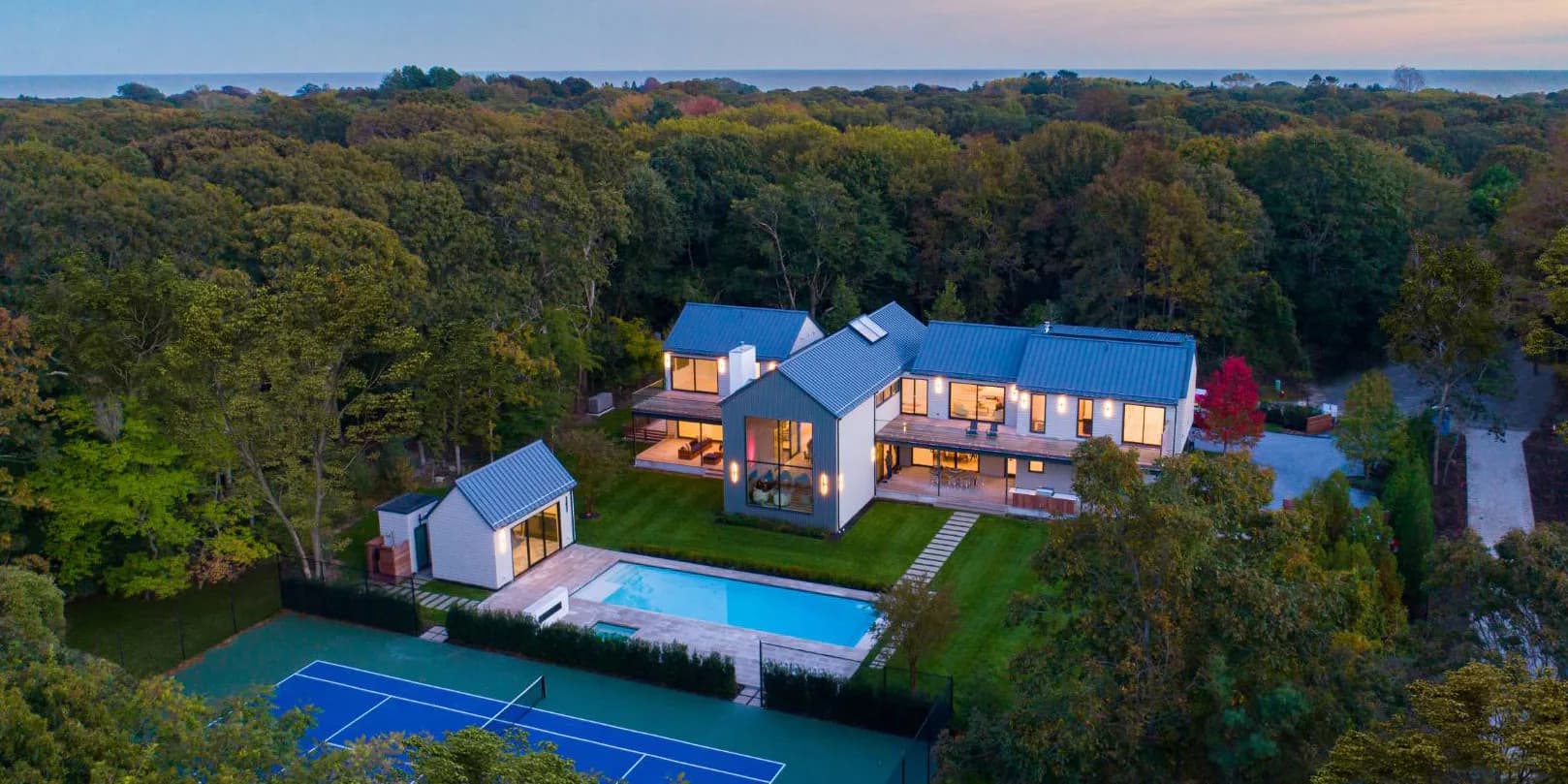 Modern residential compound with illuminated blue-clad main house, guest pavilion, and pool overlooking manicured grounds and surrounding woodland by Ayhan Ozan.