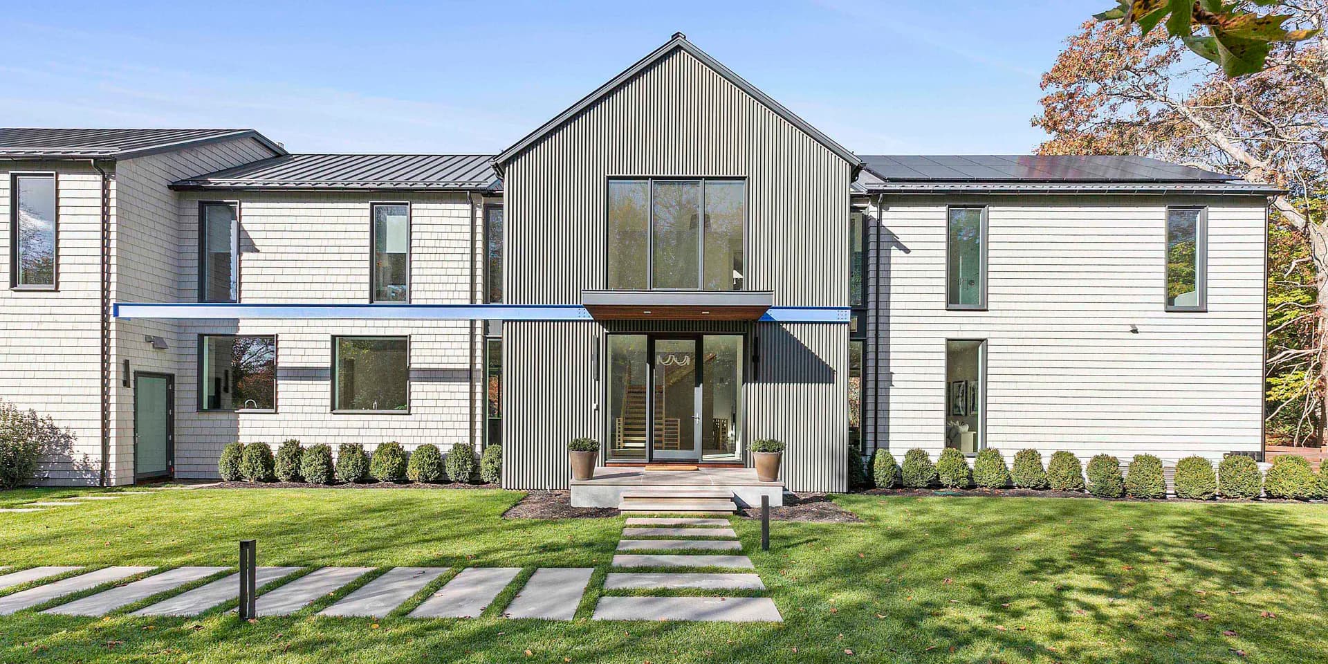 Modern residential home with mixed gray and white siding, large central gable, extensive glazing, and manicured lawn with linear plantings by Ayhan Ozan.