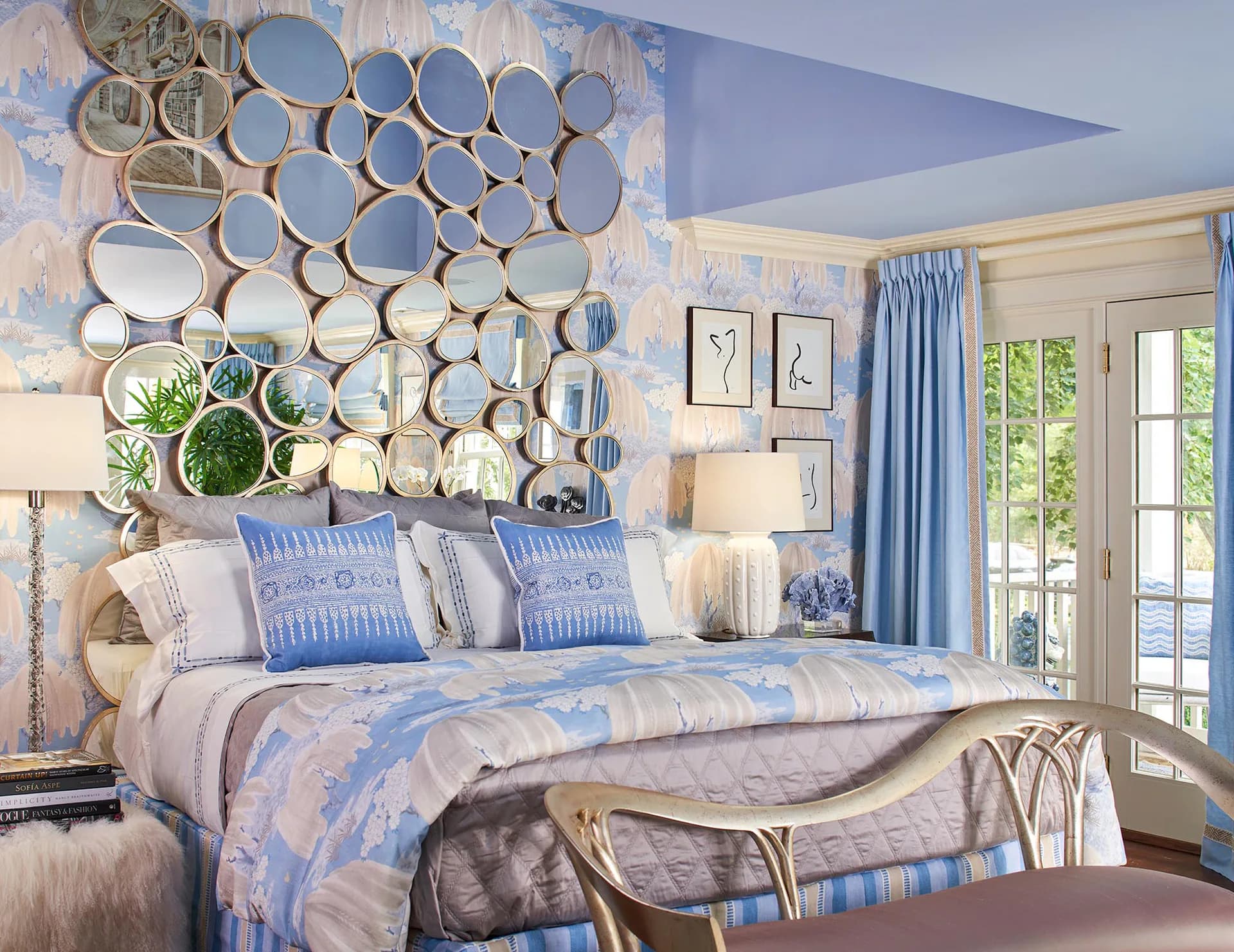 Luxurious bedroom with blue and lavender palette, circular mirror wall installation above bed, blue patterned bedding, and glass doors opening to garden by Barbara Ostrom.