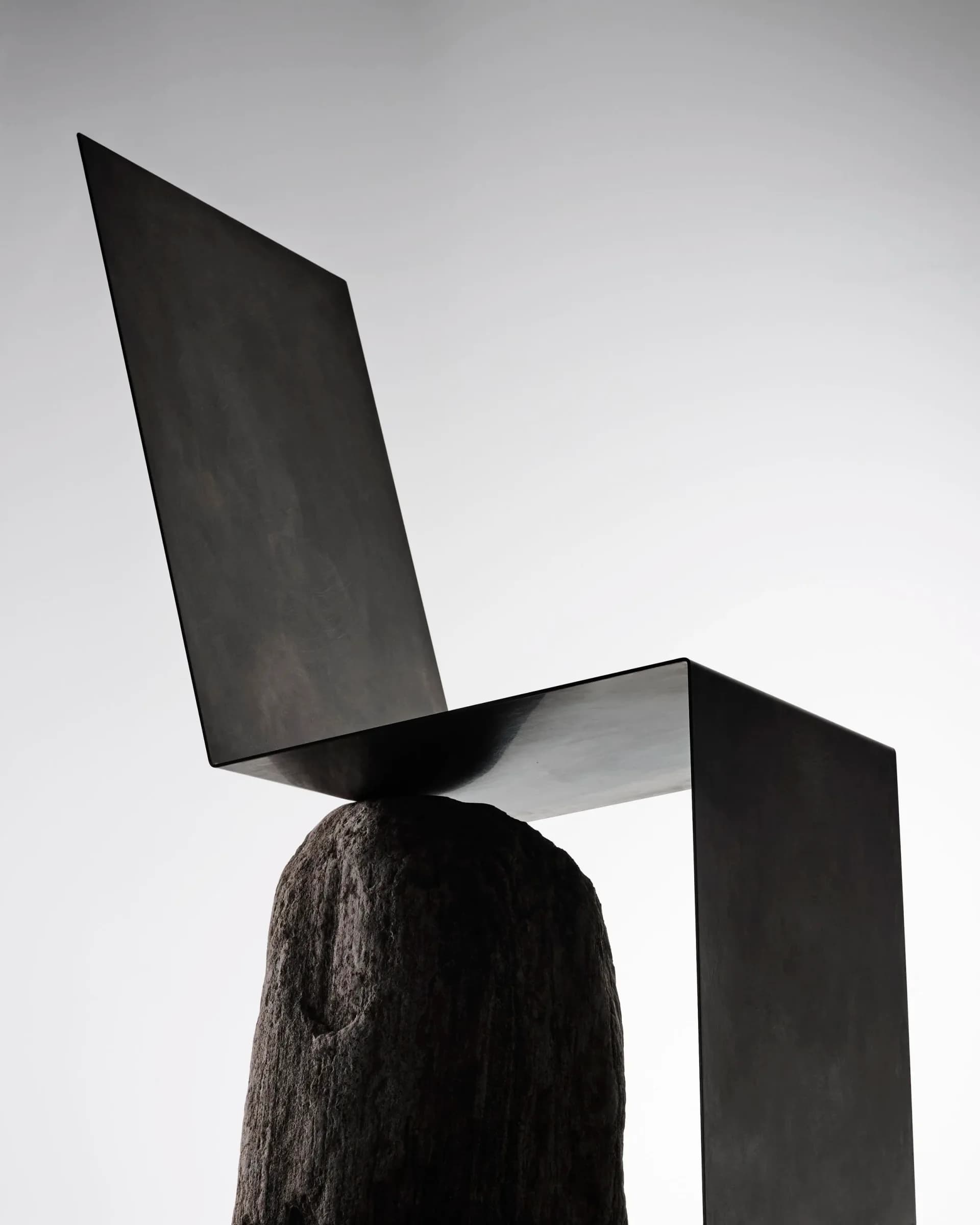 Sculptural installation featuring stacked black geometric forms—angled planes and rectangular volumes—balanced on a textured dark wooden base against a neutral background by Batten and Kamp.