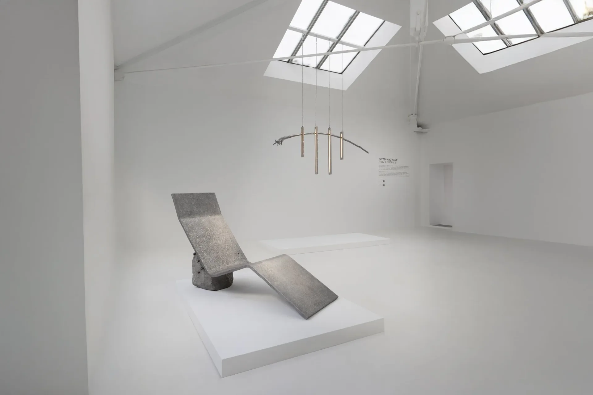 Minimalist gallery space with skylight and concrete sculptural form on white platform, featuring suspended metal fixture by Batten and Kamp.