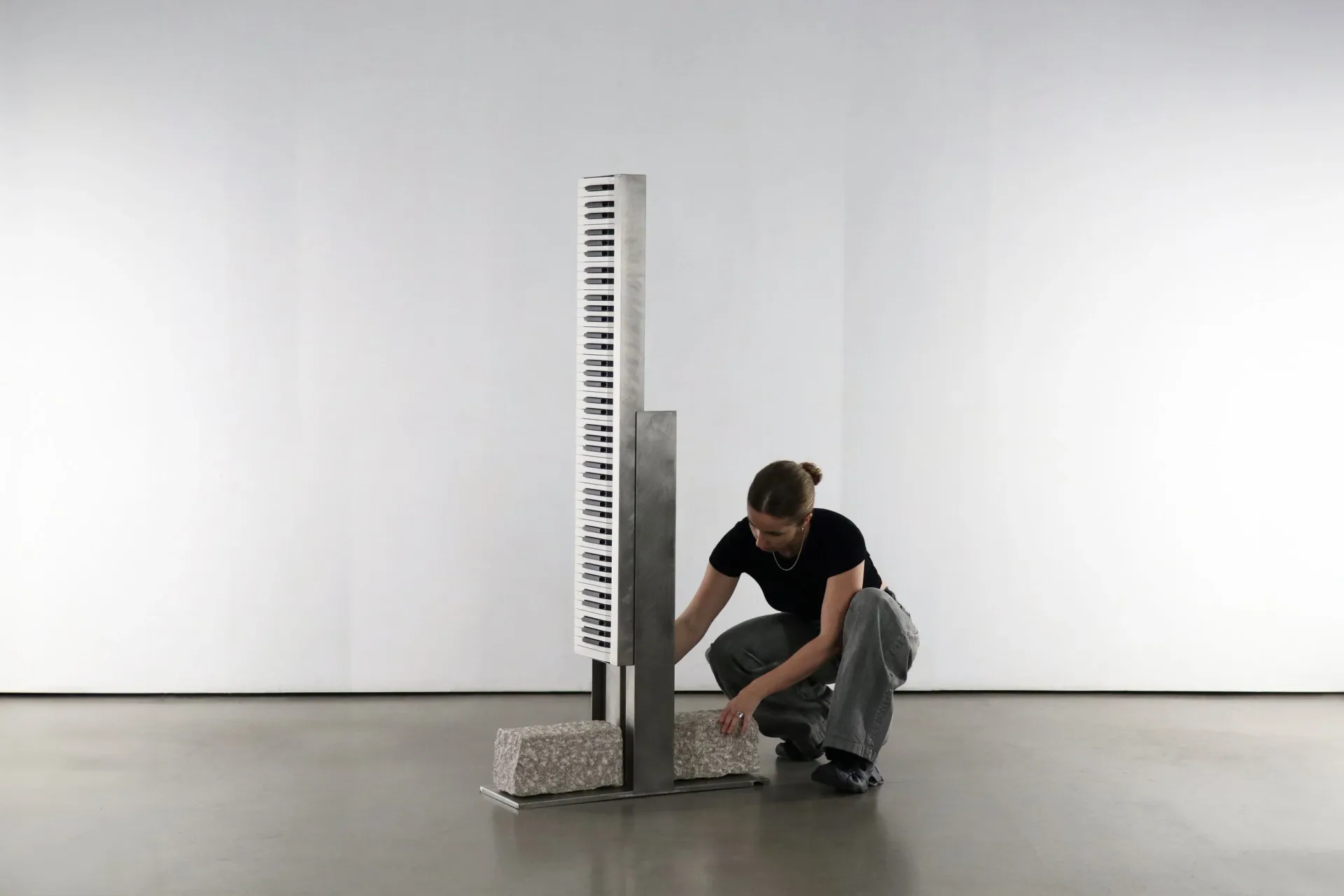 A person crouches beside a tall, slender architectural installation with horizontal ribbed detailing in a minimalist white gallery space, by Batten and Kamp.