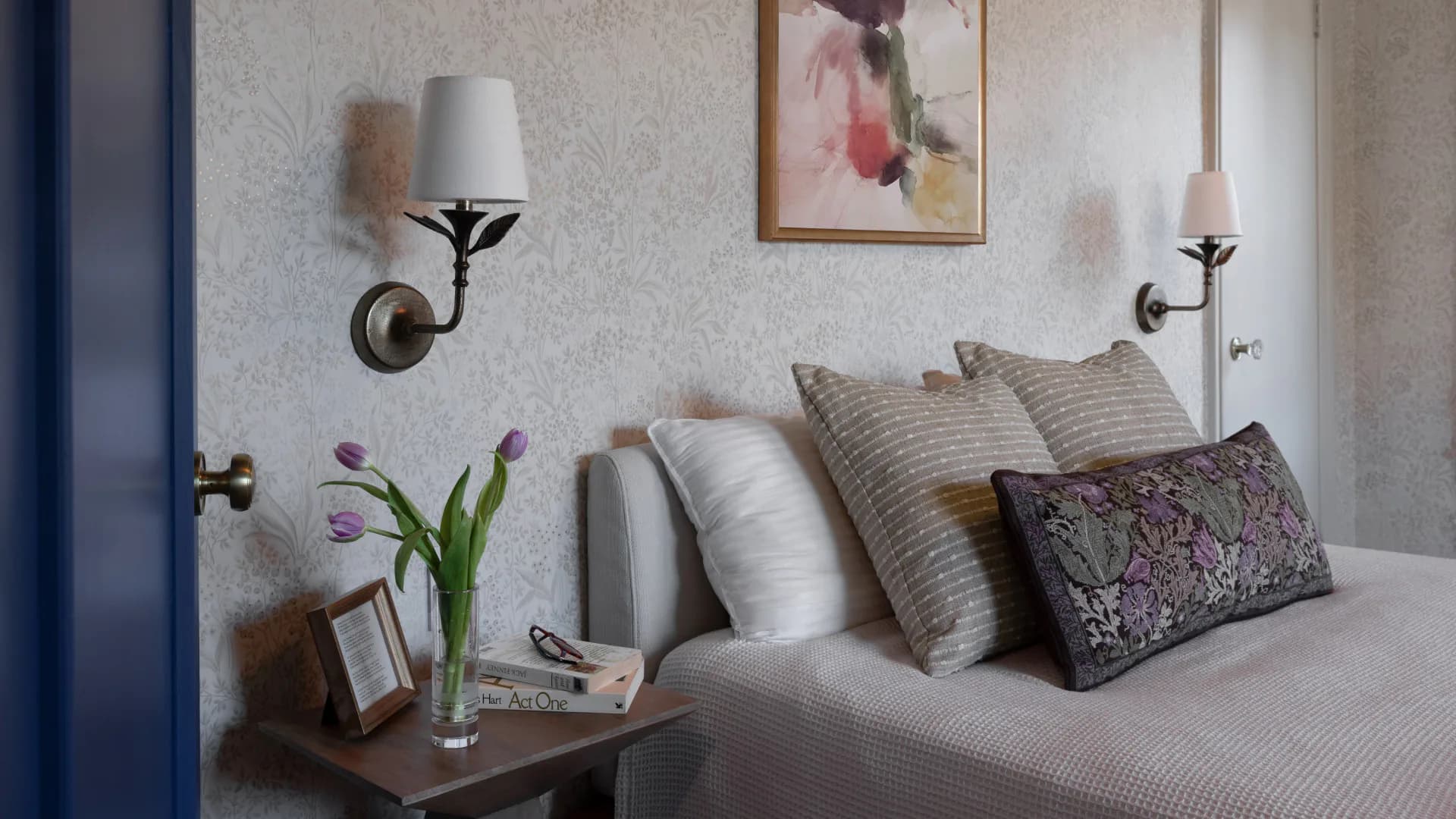 Contemporary bedroom with neutral palette featuring a bed with layered pillows, brass wall sconces, wooden nightstand with flowers, and soft textured wall finishes by Behin Forghanifar.