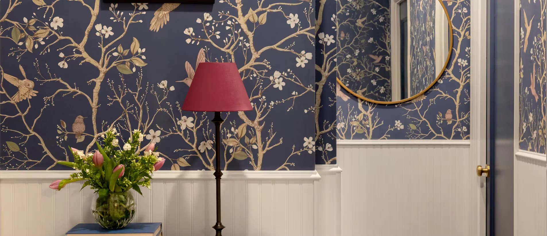 Elegant entryway featuring dark navy wallpaper with gold botanical patterns, white wainscoting, red pendant lamp, fresh green floral arrangement, and arched doorway by Behin Forghanifar.