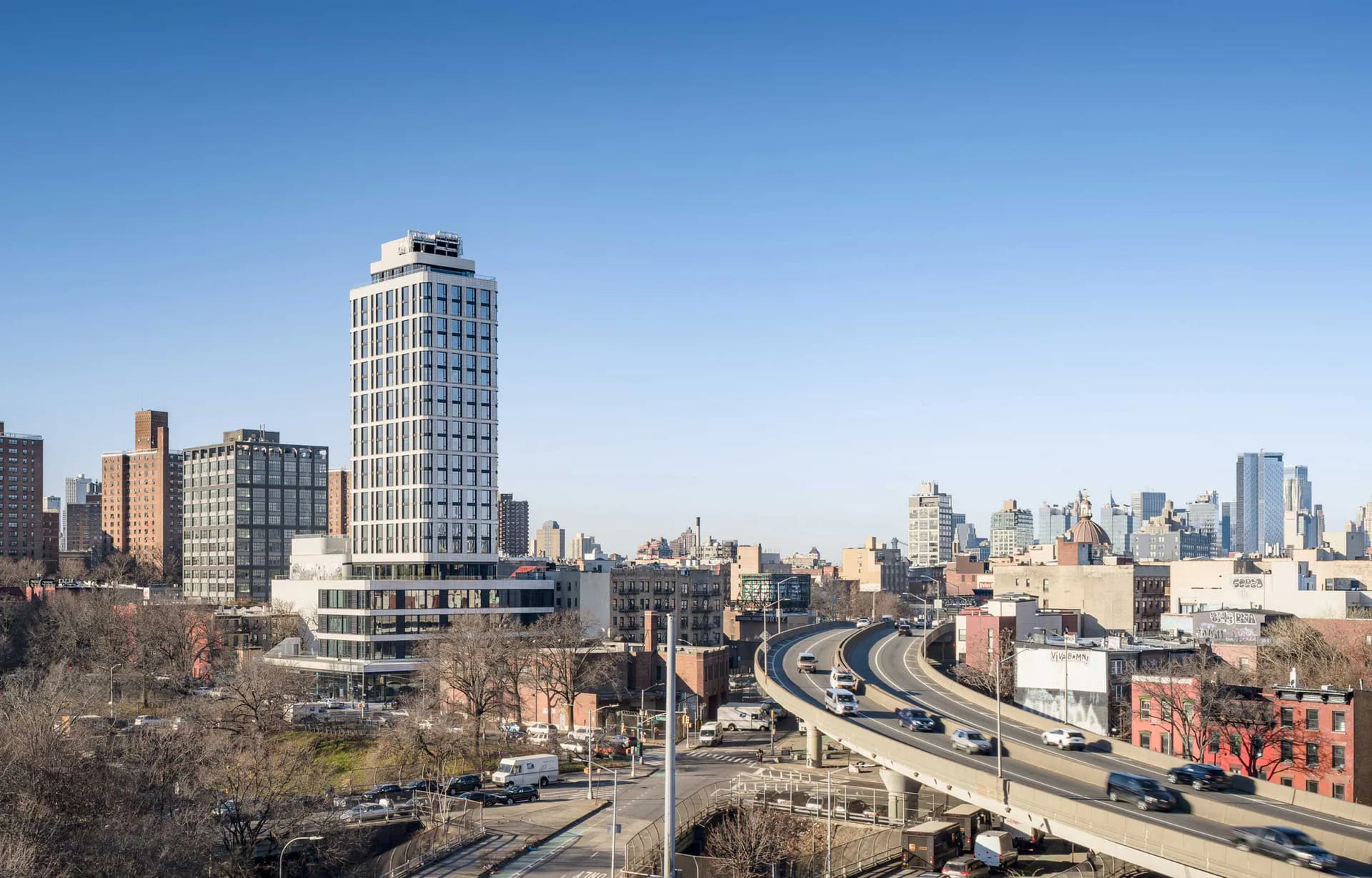 Urban cityscape featuring a tall residential tower amid mid-rise buildings and elevated highway infrastructure in New York by Bella Eastham.