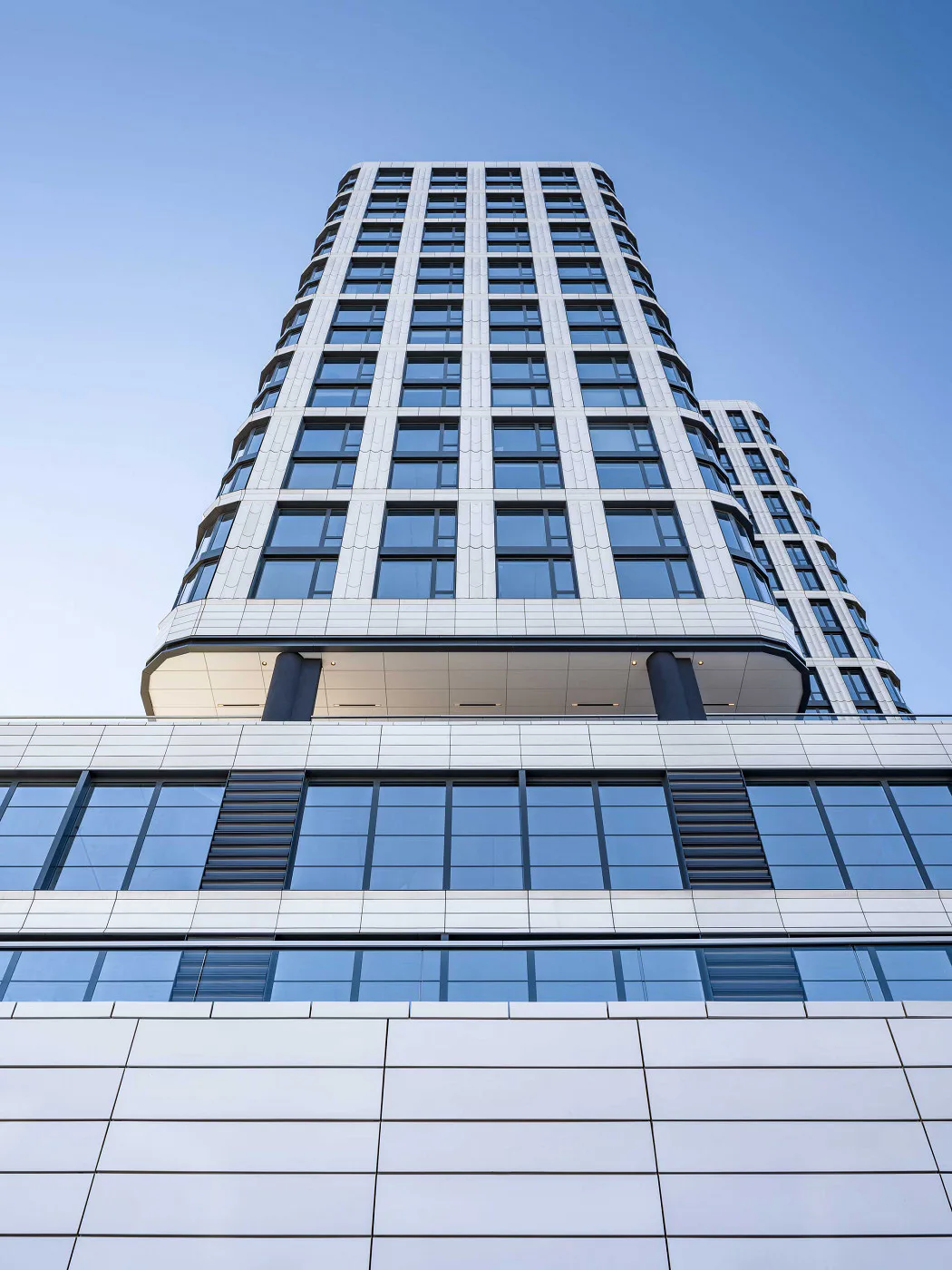 Modern curved residential tower with white and blue facade, featuring grid of windows and horizontal glass bands, photographed against clear sky by Bella Eastham.