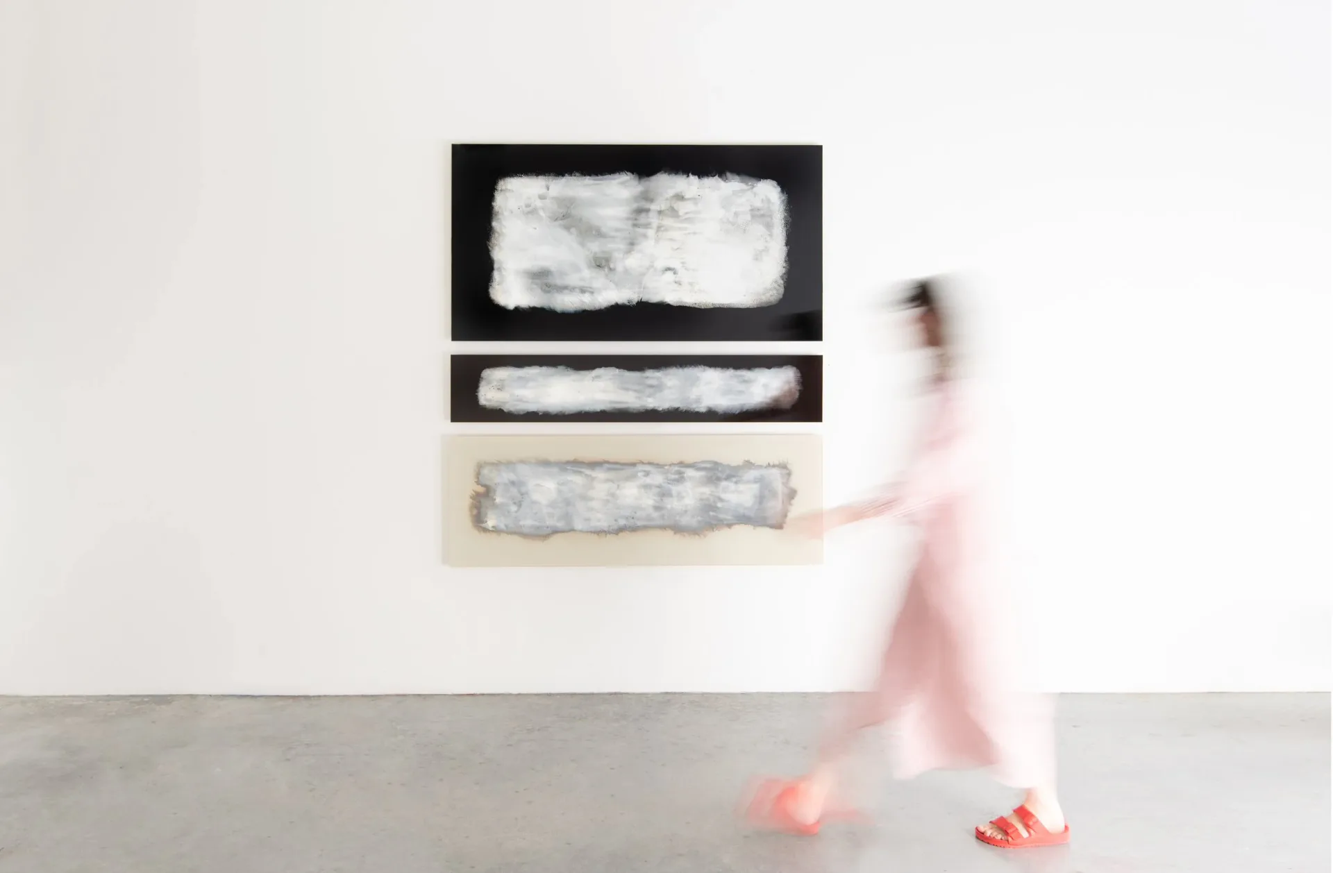 Minimalist wall installation featuring three horizontal rectangular elements with textured surfaces in black and neutral tones, photographed with a blurred figure in pink passing by, by Ben & Aja Blanc.