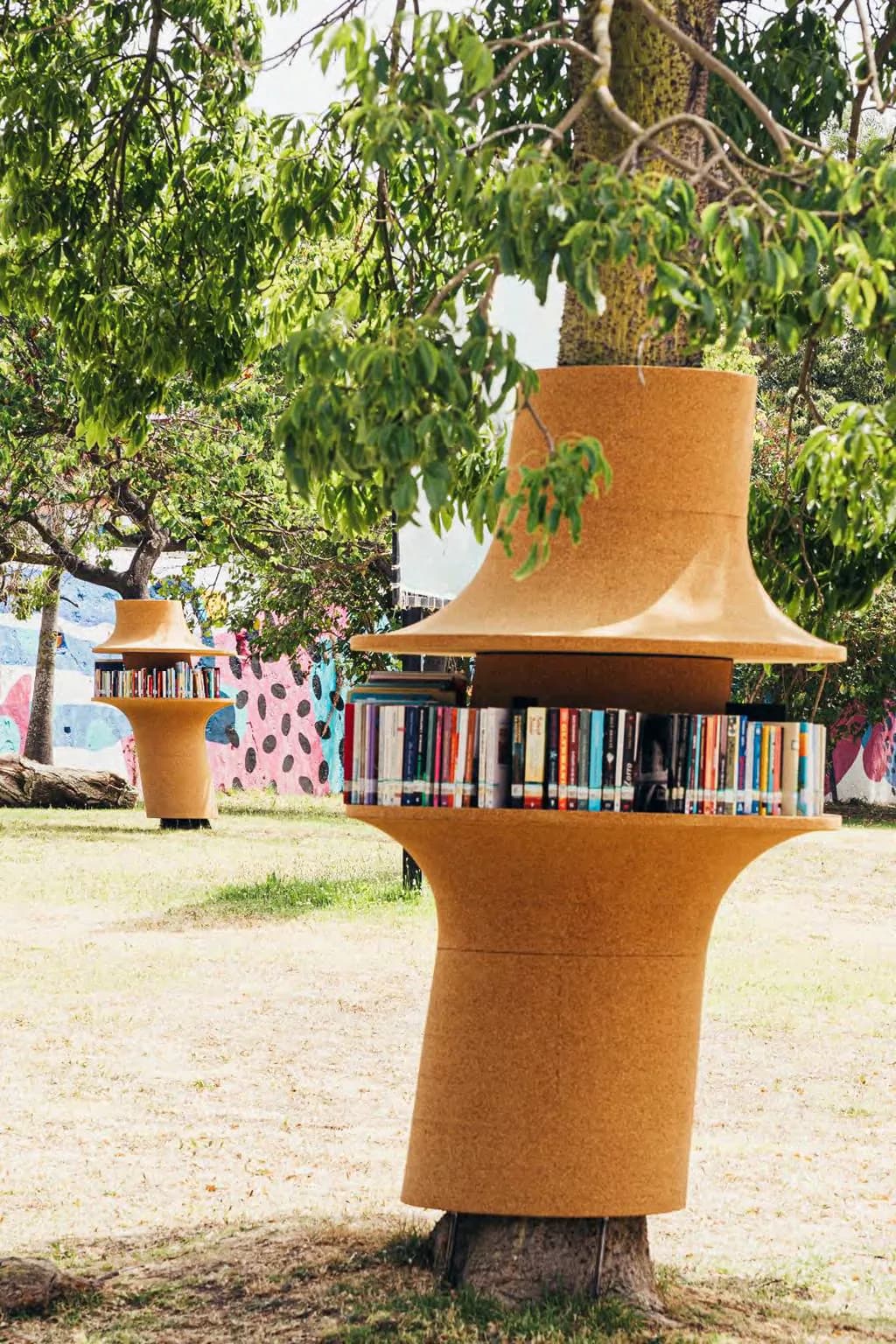 Cork cylindrical library structure with shelves of books nestled beneath trees in an outdoor park setting by Benjamin Gilmartin.