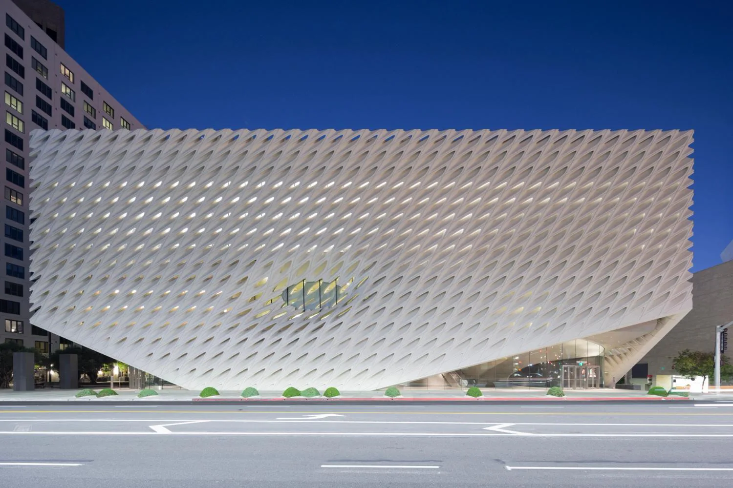 Modern white perforated facade building with geometric diagonal pattern and green interior elements, set against clear blue sky by Benjamin Gilmartin.