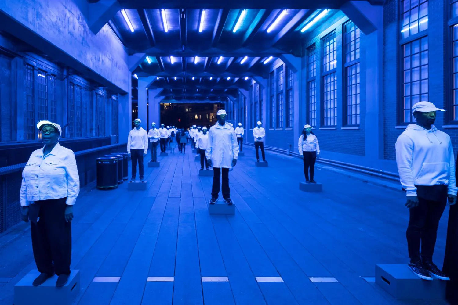 A blue-toned corridor with synthetic figures in white clothing spaced throughout, featuring linear ceiling fixtures and geometric flooring—a digital rendering by Benjamin Gilmartin.