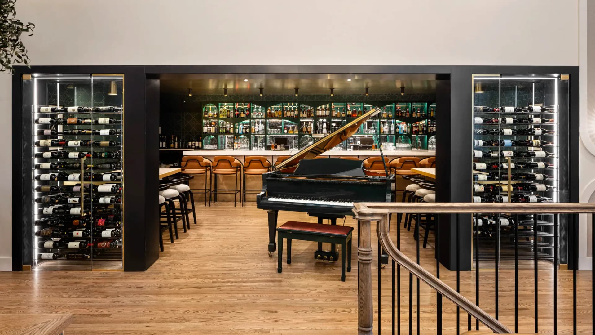 A sophisticated wine bar and lounge features a grand piano, wooden flooring, dark cabinetry with wine storage on either side, and a stocked bar counter in warm wood tones by Beth Donner.