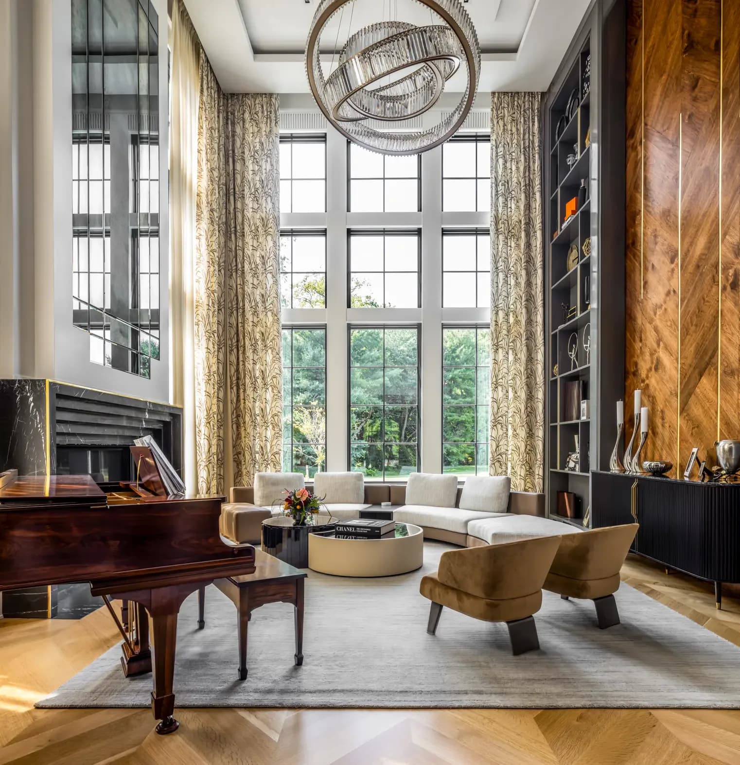 Luxurious double-height living room with floor-to-ceiling windows, grand piano, brass-framed chairs, floating circular pendant light, and wood paneling by Beth Donner.