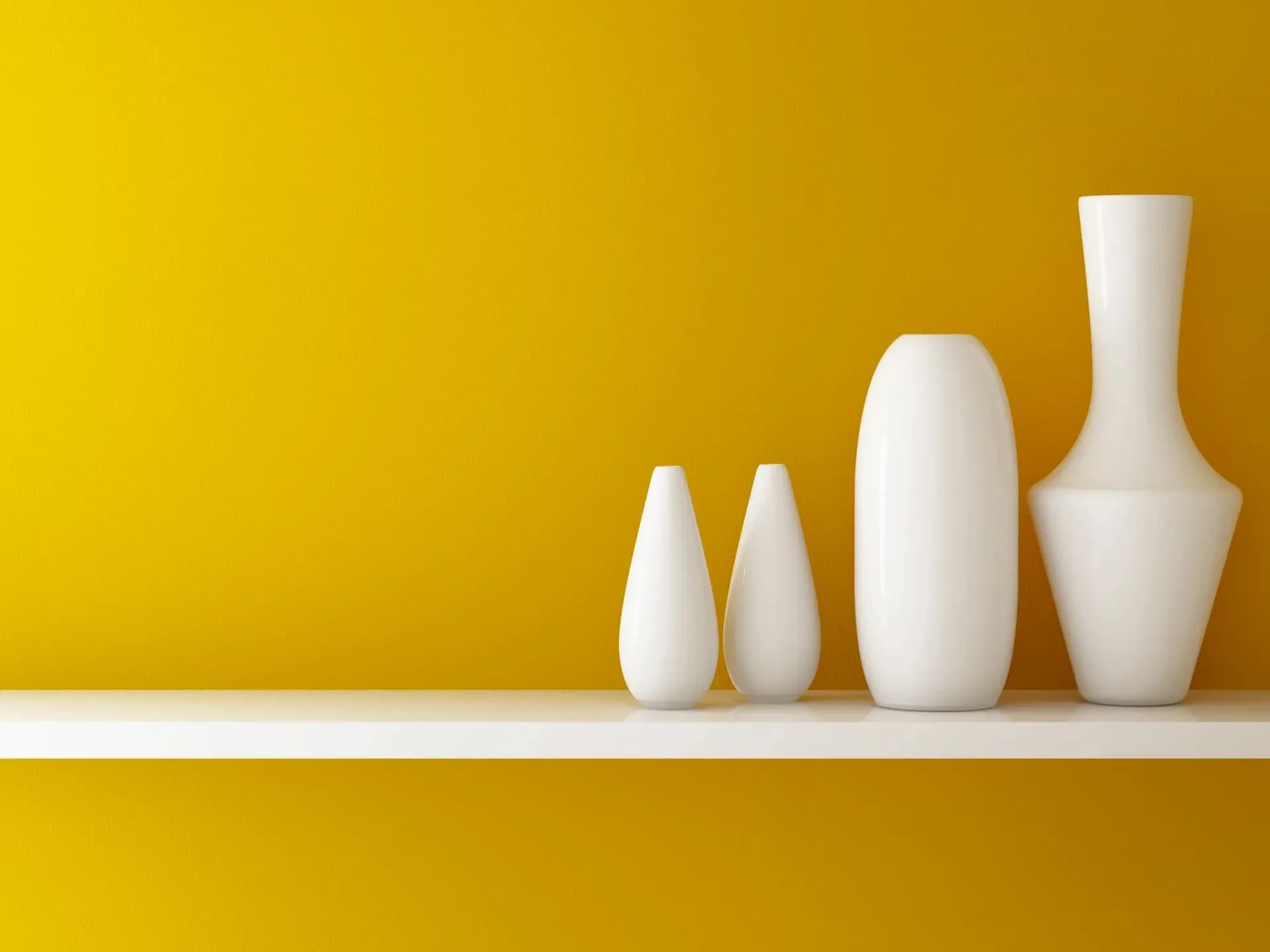 White ceramic vases of varying heights arranged on a white shelf against a yellow wall by Beth Freeberg.