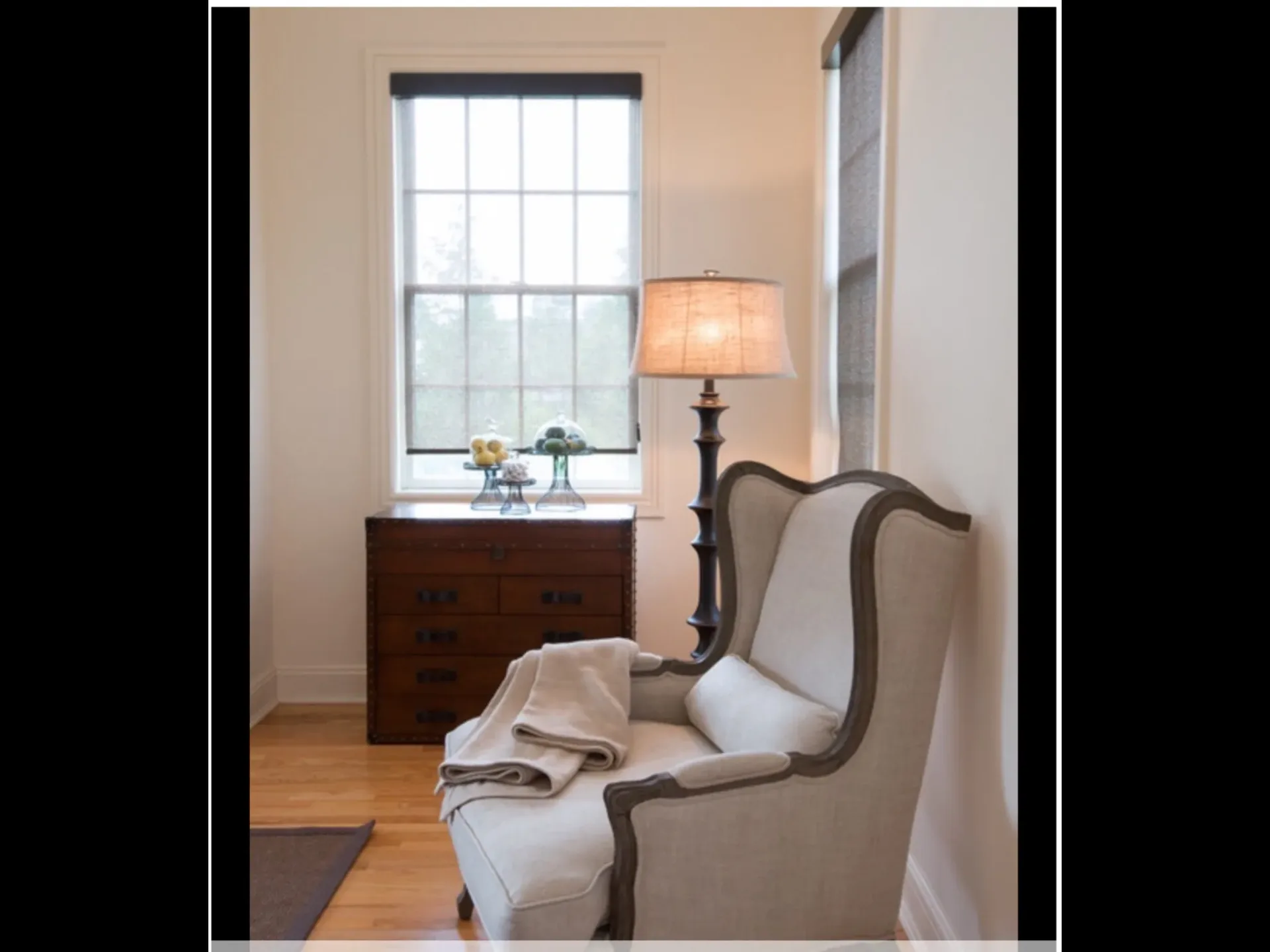 Elegant sitting area with upholstered wingback chair, wooden dresser, table lamp, and large window with neutral palette by Beth Freeberg.