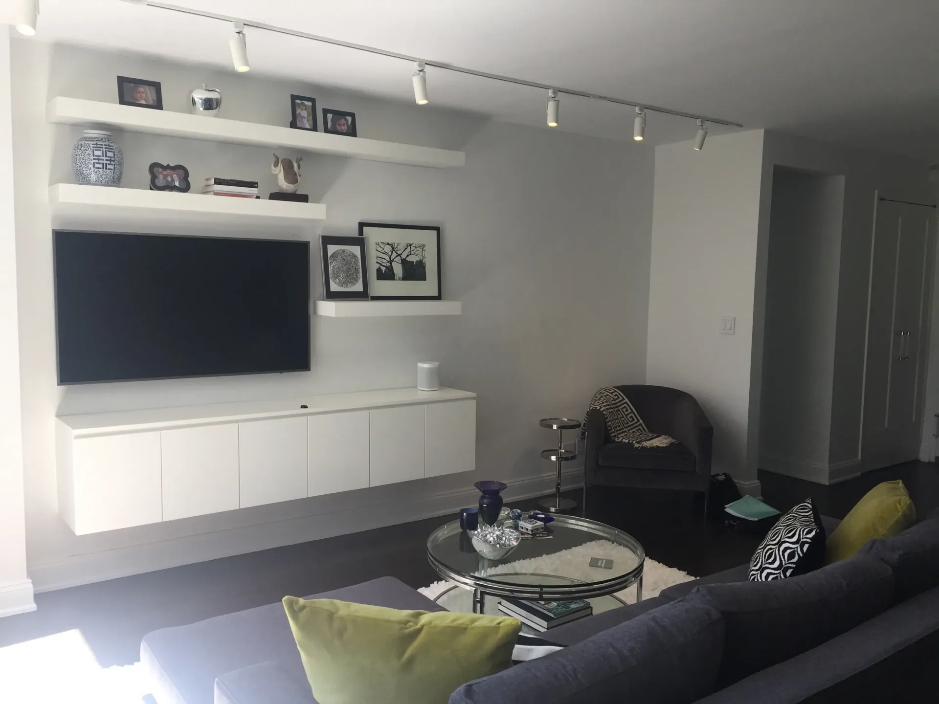 Modern living room with dark gray sofa, yellow-green accent pillows, white wall-mounted media console with TV, floating shelves, and round glass coffee table by Beth Freeberg.