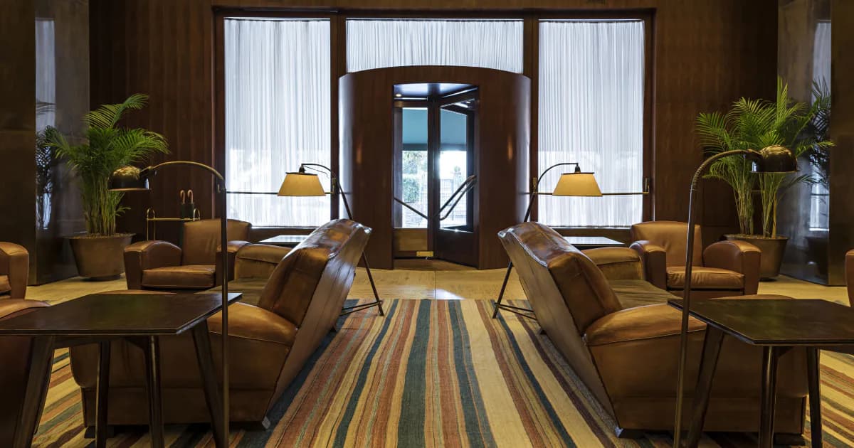 Luxury hotel lobby with warm wood tones, striped carpet runner, symmetrical seating areas with leather chairs, and arched doorway with natural light by Bianca Salles Bia.