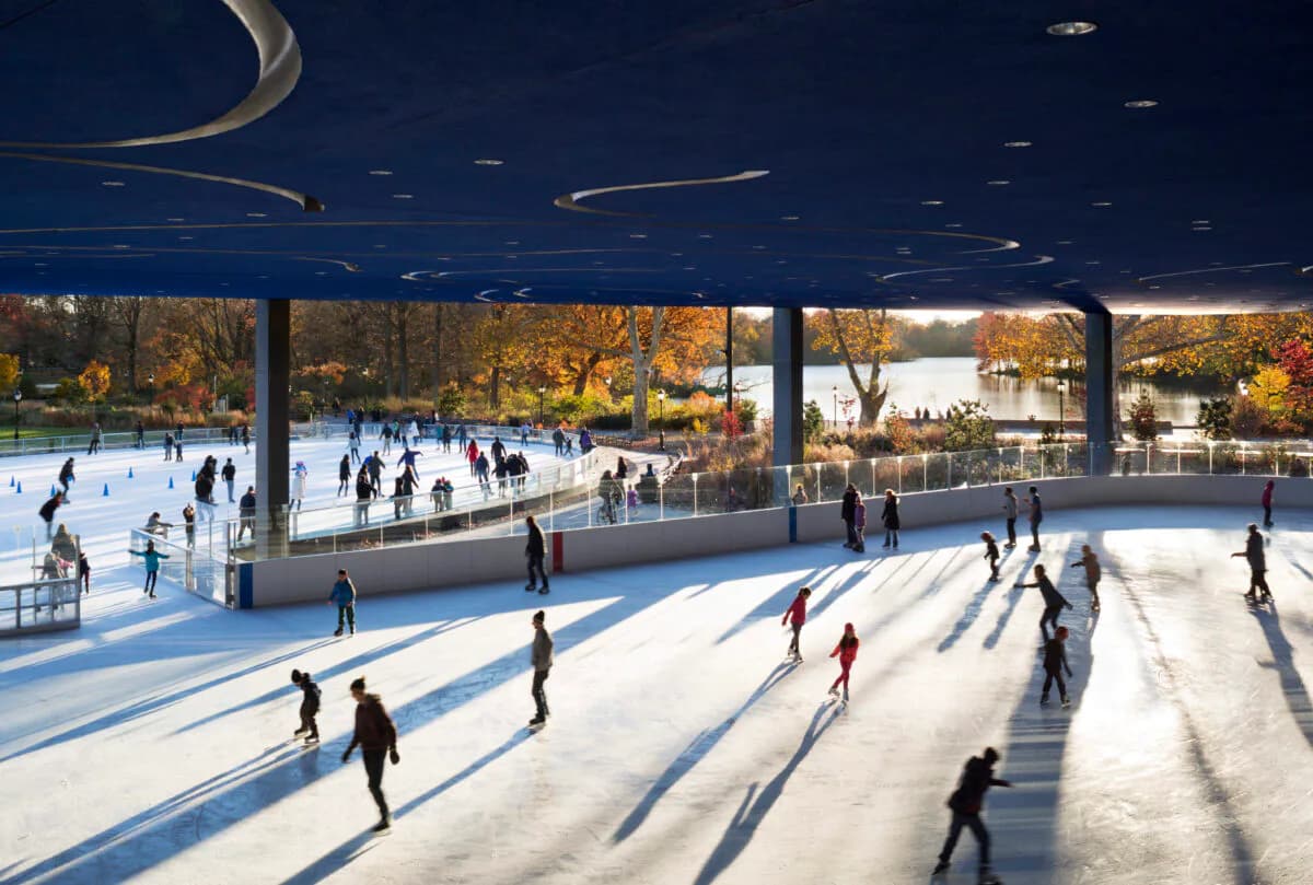 Modern ice skating rink with expansive glazing overlooking autumn trees, featuring dramatic ceiling details and multiple skaters casting long shadows across the blue ice surface by Billie Tsien.