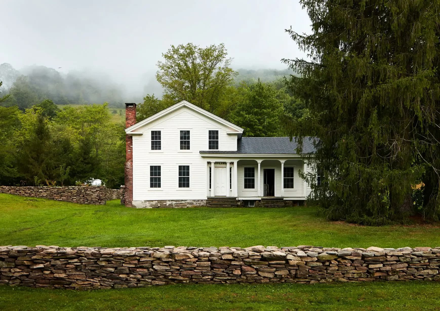 Classic white farmhouse with brick chimney set on manicured green lawn, framed by stone wall and evergreens, mountains visible in misty background, by Billy Cotton.